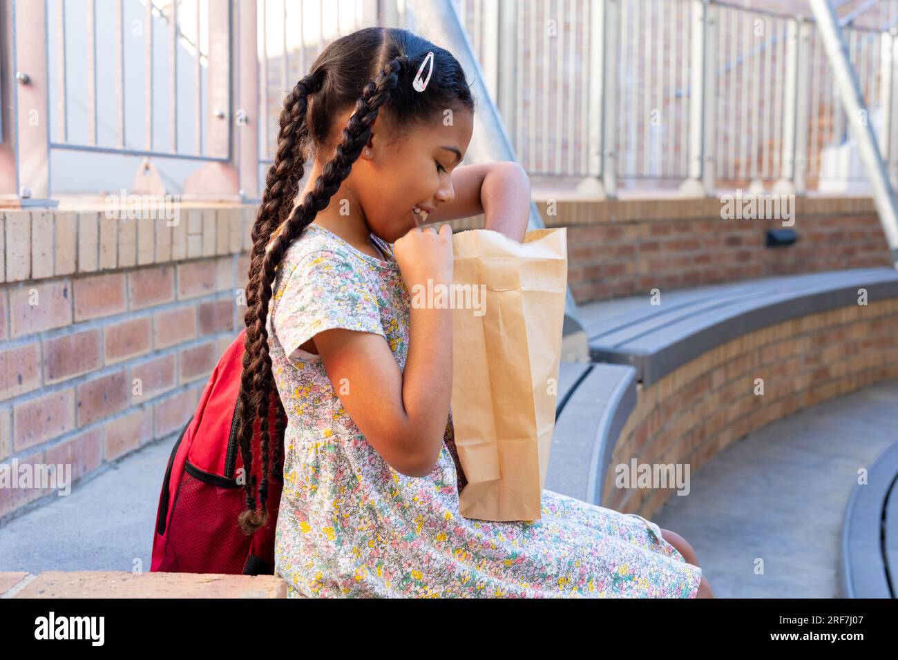 Felice studentessa birazziale seduto sulla panchina e aperto il pranzo fuori dalla scuola elementare Foto Stock