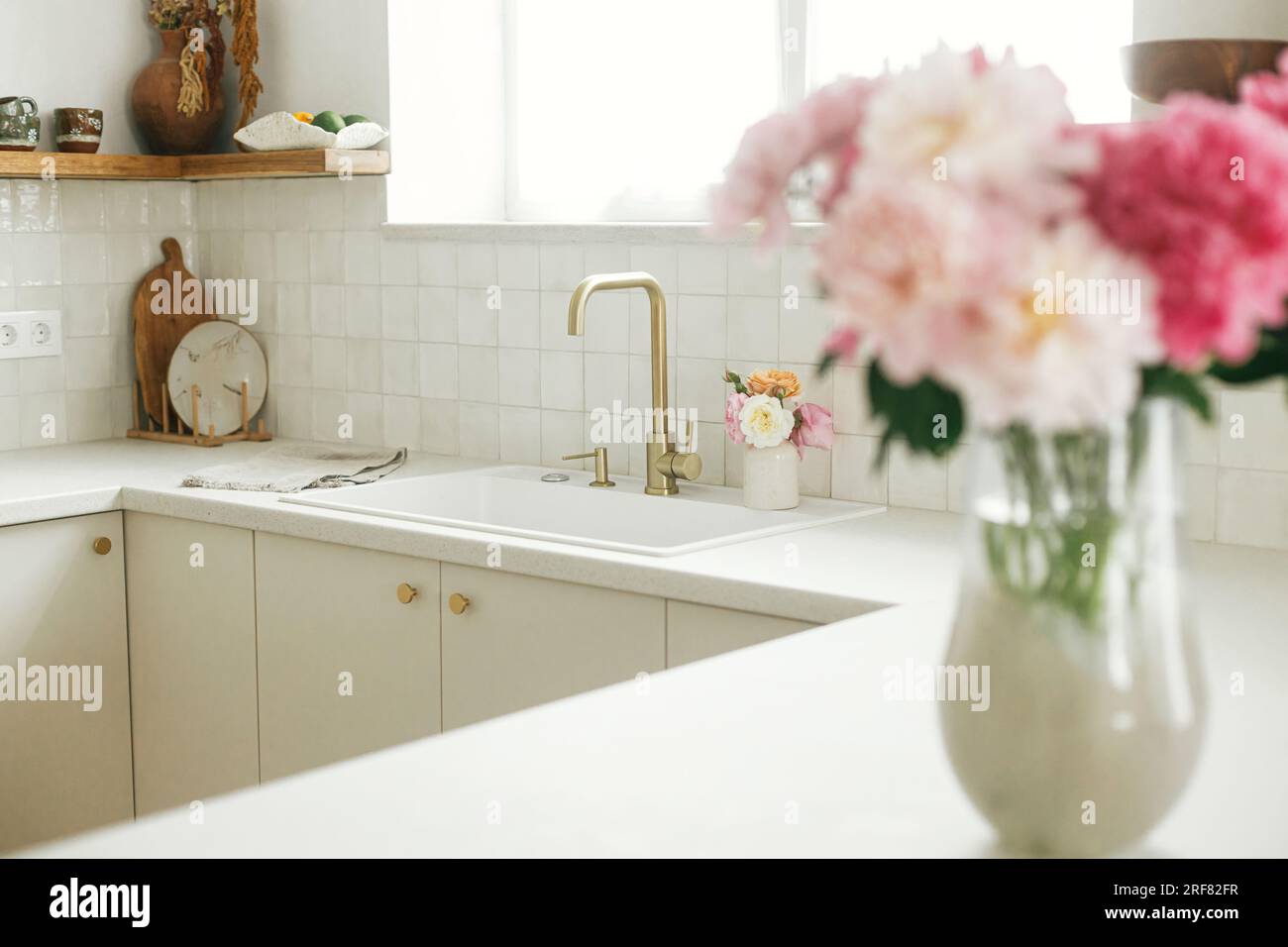 Cucina dal design moderno. Elegante rubinetto in ottone e lavandino in granito bianco, armadietti da cucina con pomelli e bouquet di peonie nella nuova casa scandinava. Moderno Foto Stock