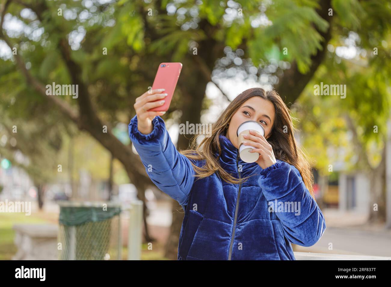 Ragazza latina con una tazza di caffè usa e getta che scatta un selfie con il cellulare in un parco pubblico. Foto Stock