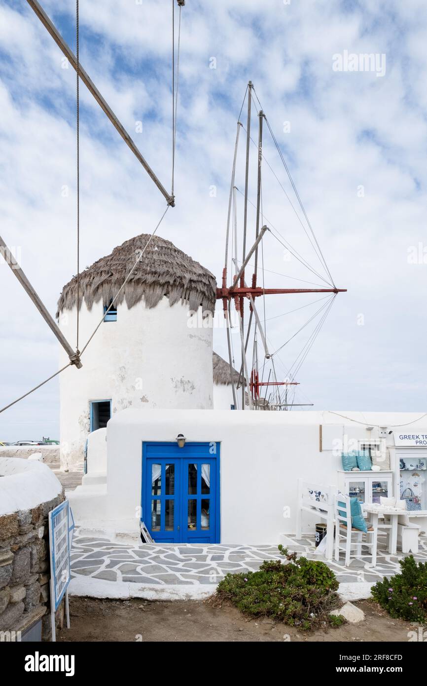 Mulini a vento di Mykonos dal lato, con un piccolo edificio con una porta con cornice blu davanti e cielo blu nuvoloso sopra. Foto Stock