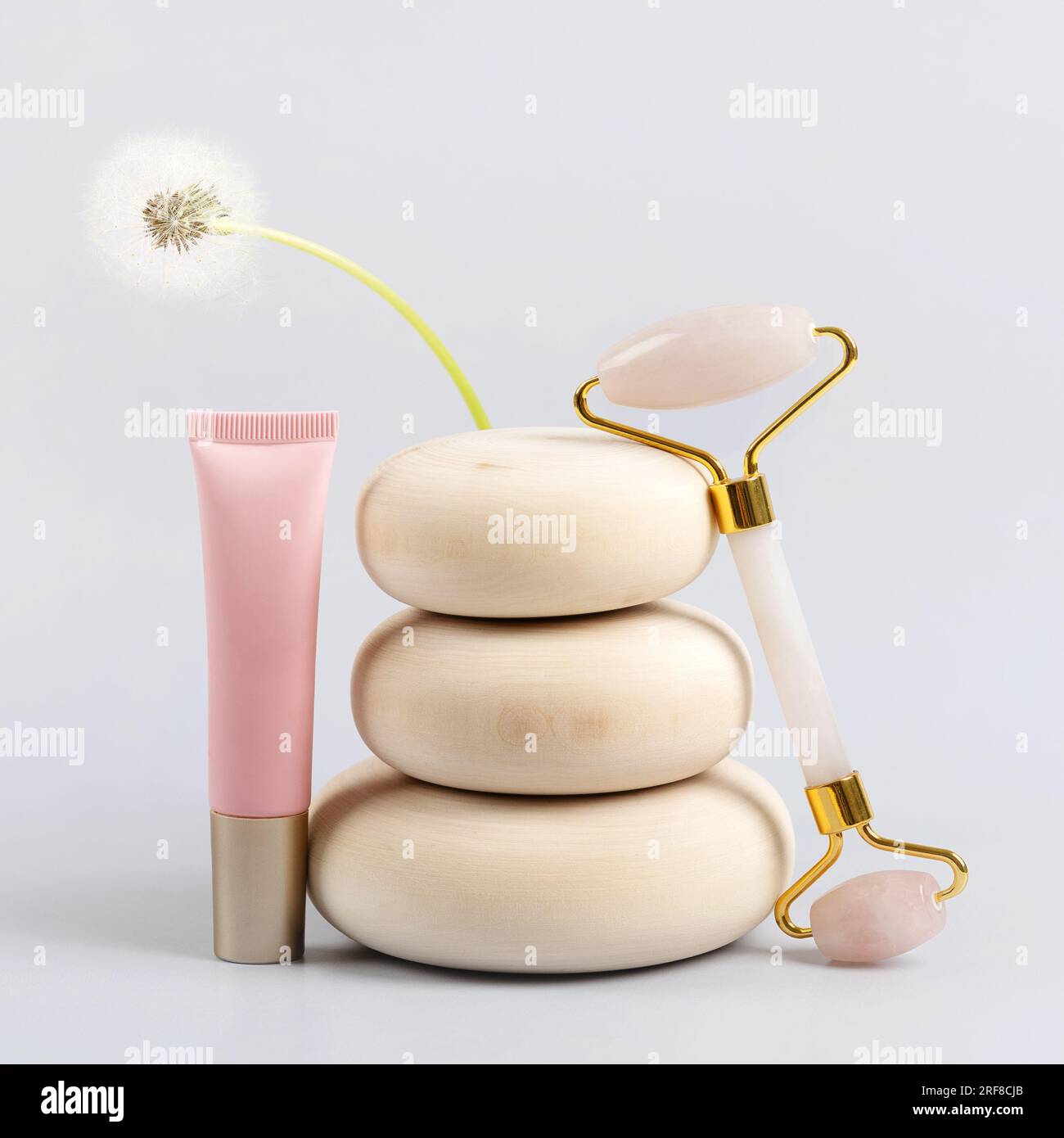 Rullo facciale in quarzo rosa e contenitore per bottiglie cosmetiche su pietre di legno con dente di leone floreale, sfondo grigio. Massaggio viso anti-età per l. Naturale Foto Stock
