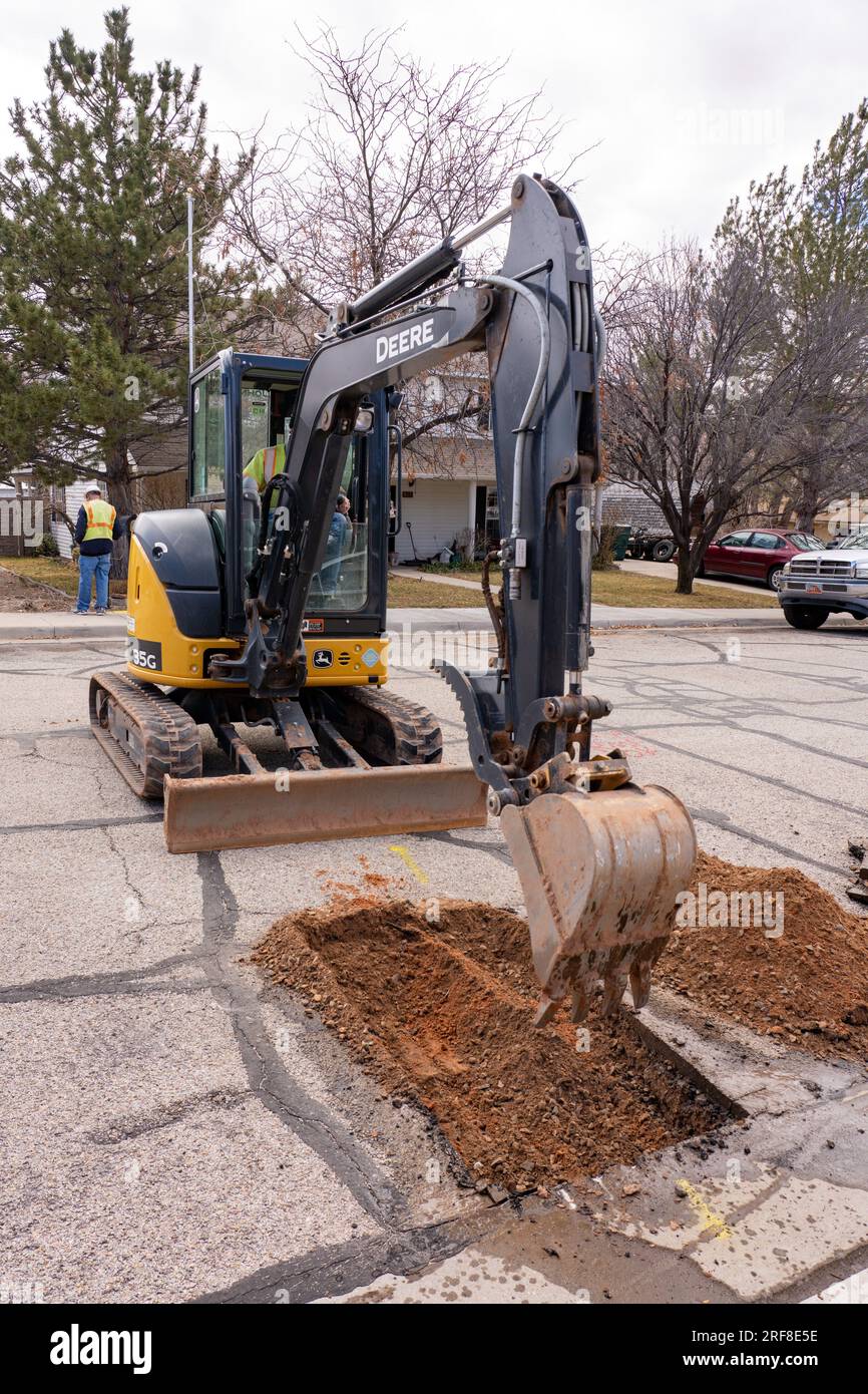 Un operaio utilizza una zappa cingolata per scavare un foro nella strada e riparare una linea di servizi pubblici in un quartiere. Foto Stock