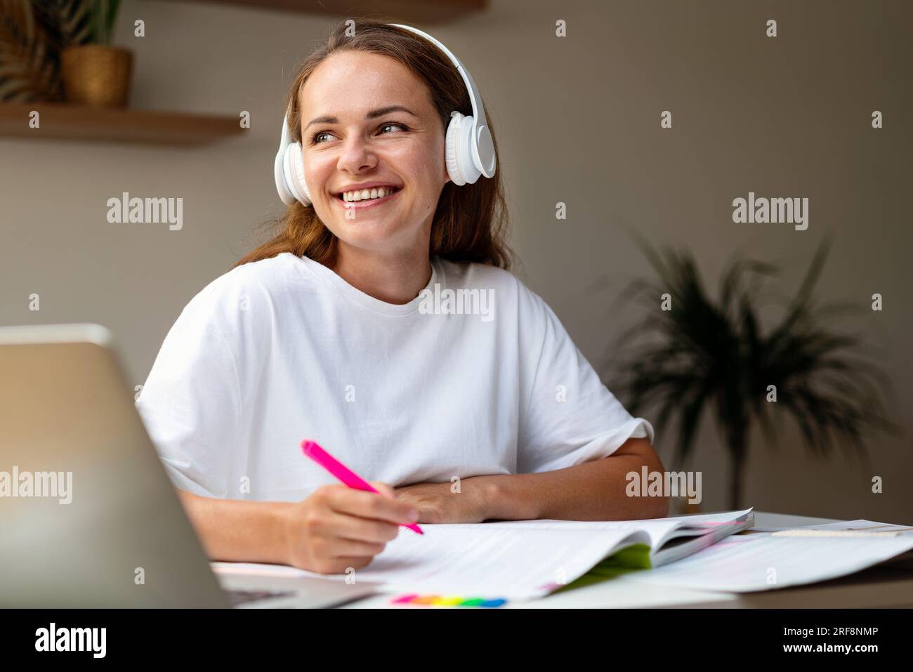 Studentessa felice in cuffie sedute alla scrivania a casa a fare i compiti nel libro degli esercizi. Foto Stock
