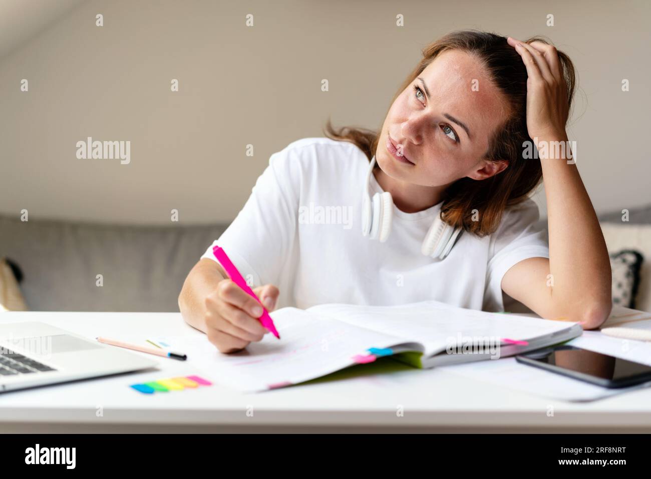 donna bruna che indossa una maglietta bianca seduta alla scrivania a casa e che studia, fa i compiti, pensa all'esercizio nel libro di testo Foto Stock