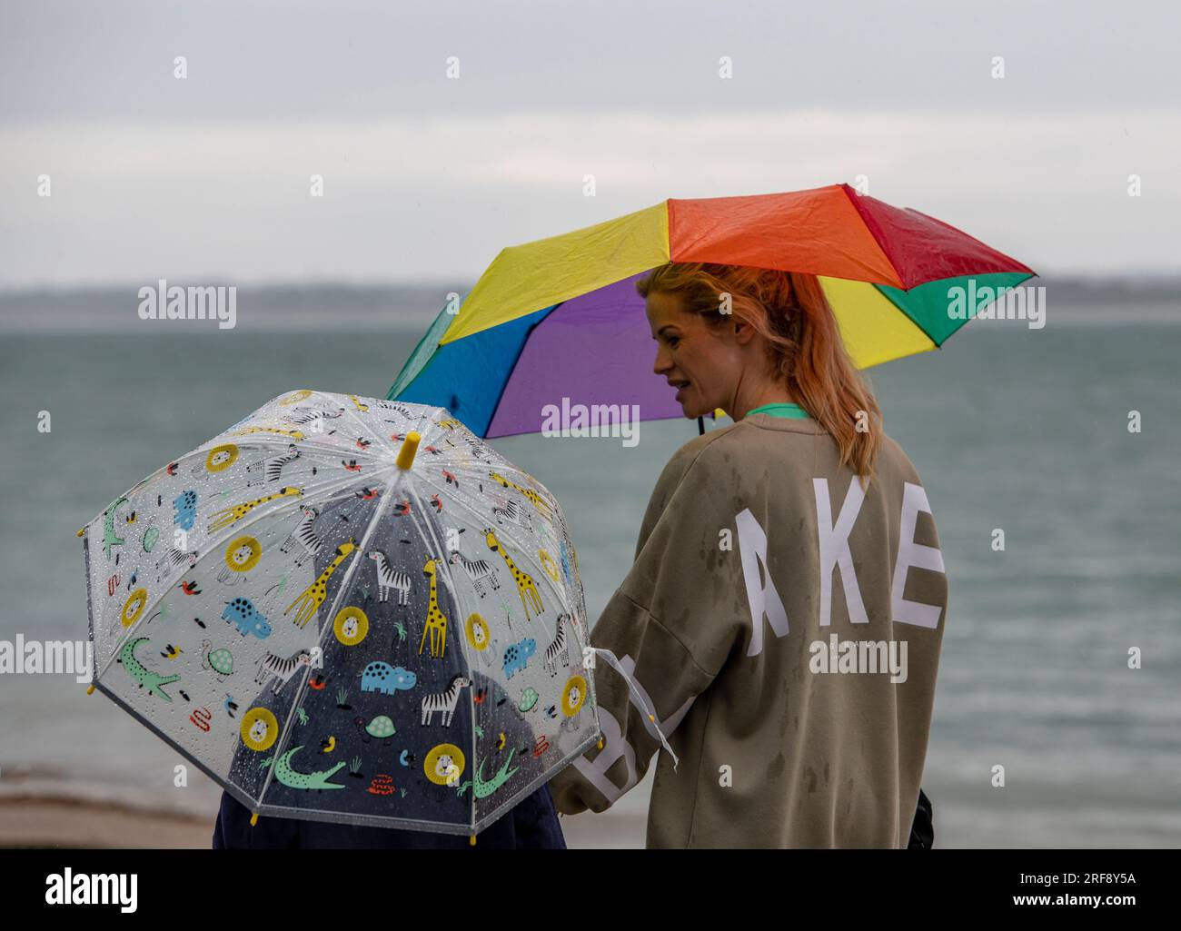 madre e figlia camminano insieme sotto la pioggia con ombrelli nuvolosi e luminosi. mamma e bambino usano ombrelli in una giornata di pioggia al mare. Foto Stock