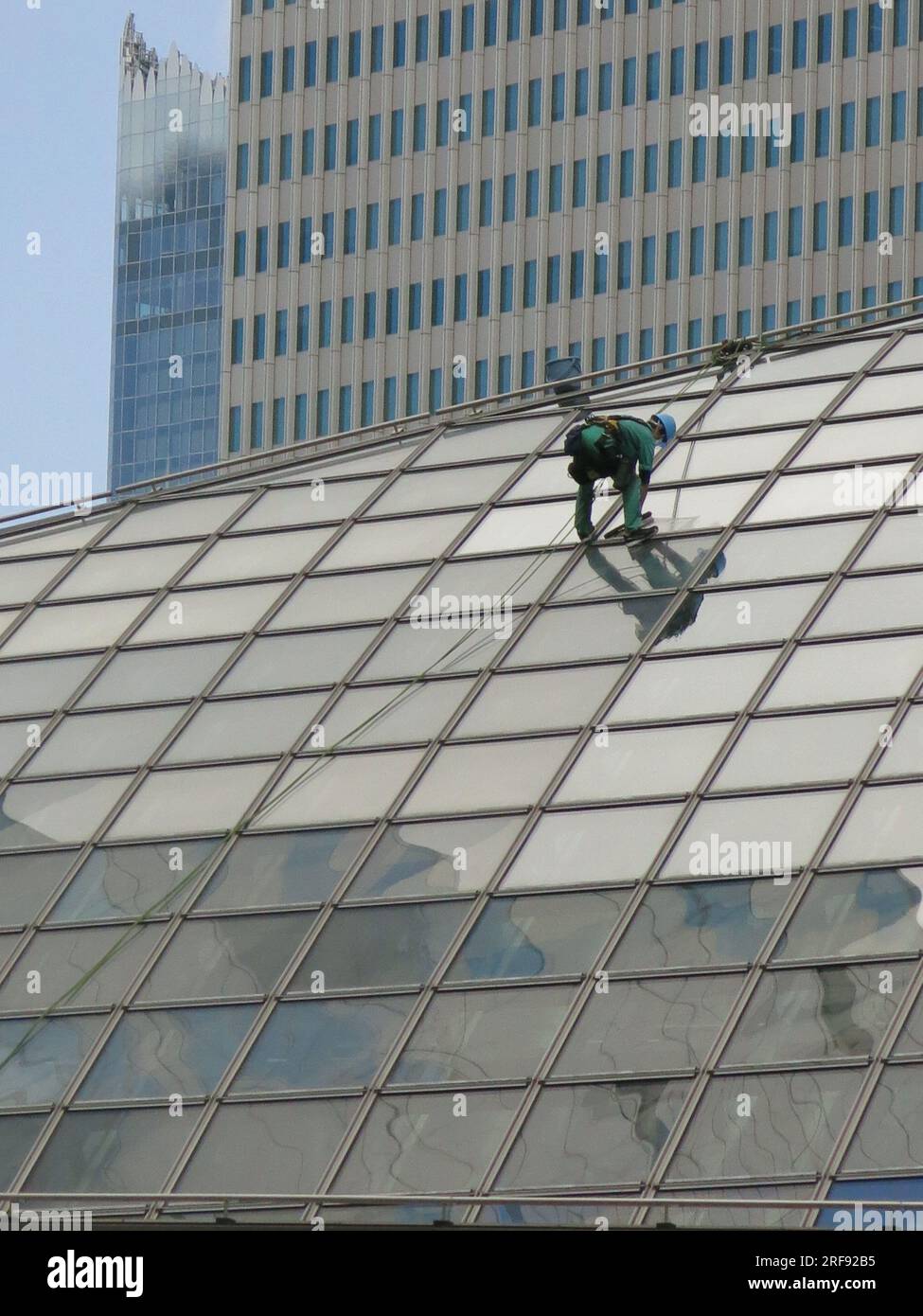 Un addetto alla pulizia dei finestrini indossa indumenti protettivi con un casco e un'imbracatura mentre è collegato a una corda su un ripido tetto di vetro di un moderno grattacielo Yokohama. Foto Stock
