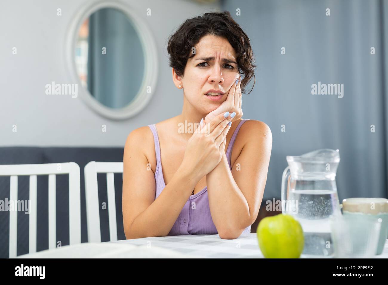 Giovane donna con mal di denti e guancia toccante, seduta a tavola Foto Stock