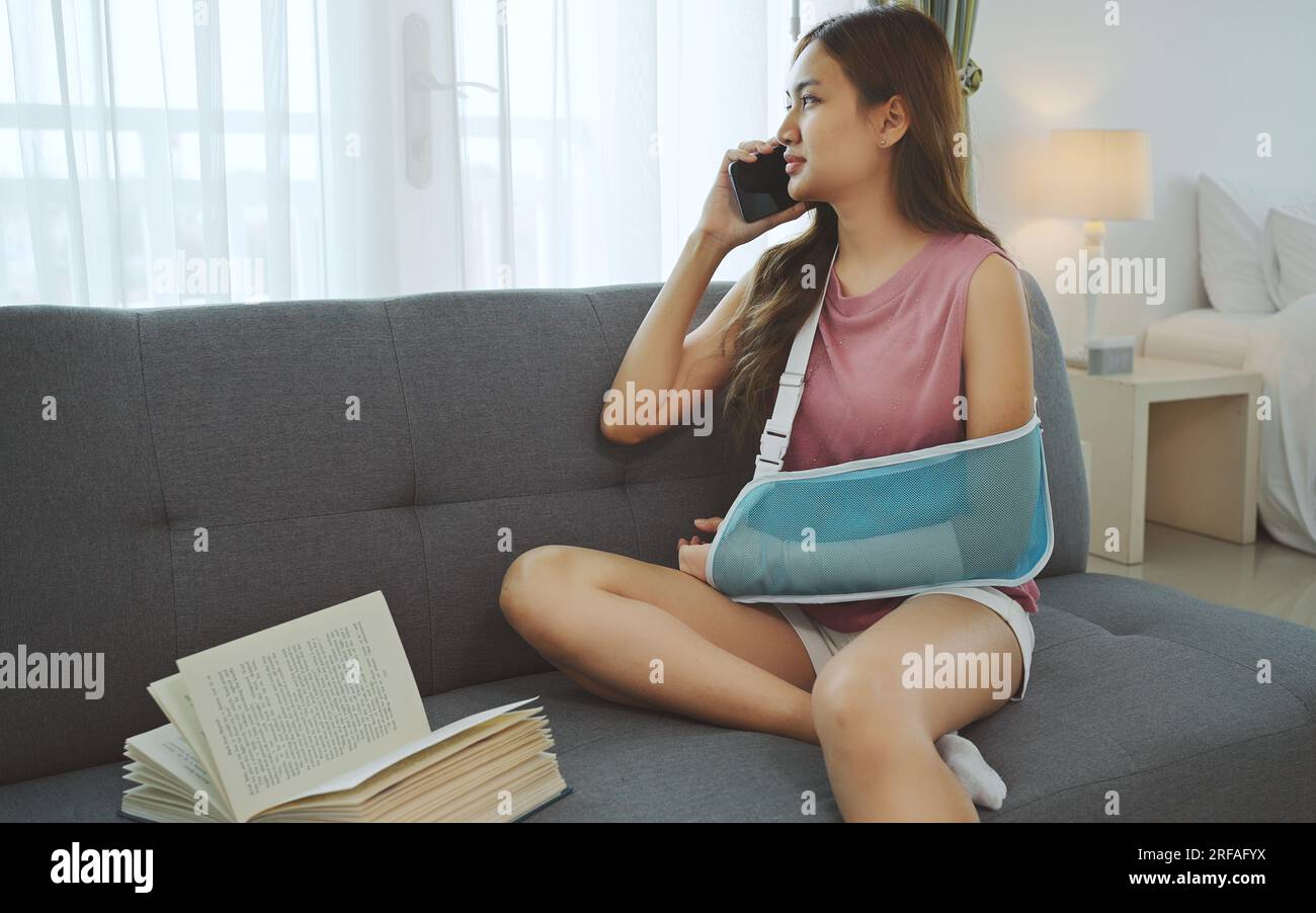Giovane donna asiatica che indossa una morbida stecca a causa del mal di braccio che usa il telefono sul divano a casa. Foto Stock