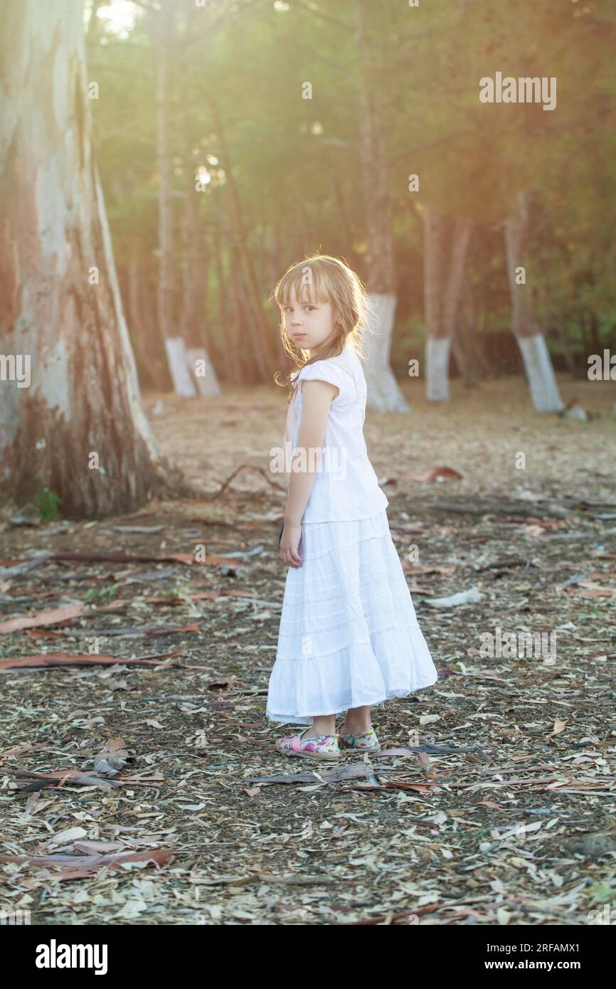Bruna bambina in abiti bianchi in piedi lateralmente sullo sfondo del parco soleggiato Foto Stock