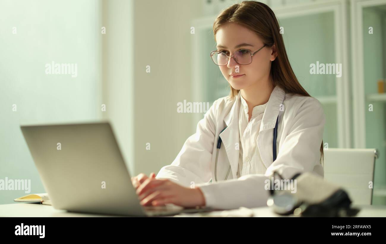 Medico femminile concentrato siede a tavolo in ospedale e lavora su portatile online Foto Stock