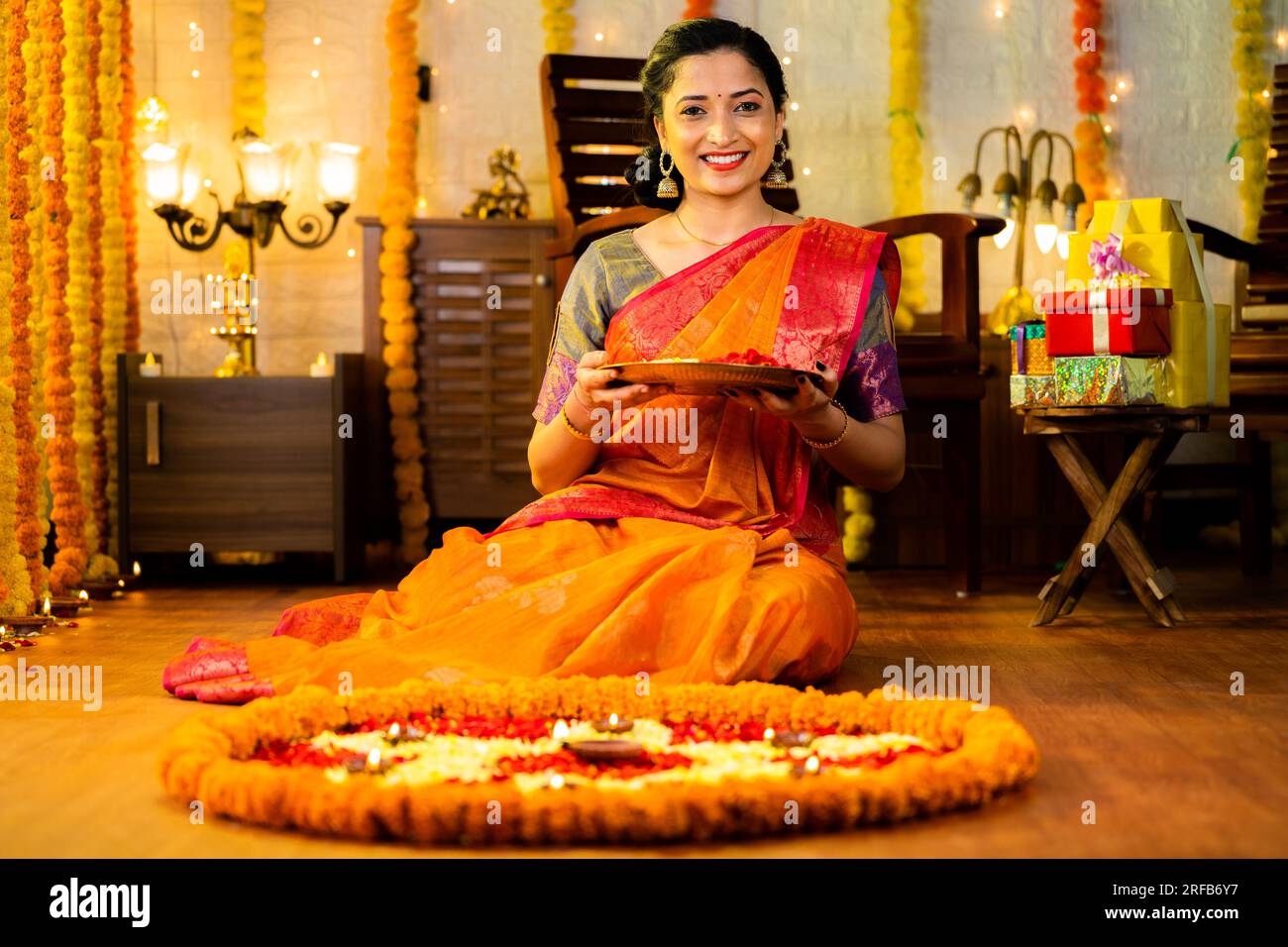Ampia ripresa di una felice ragazza indiana sorridente che tiene un piatto di fiori guardando la macchina fotografica davanti alla decorazione del pavimento - concetto di celebrazione diwali Foto Stock