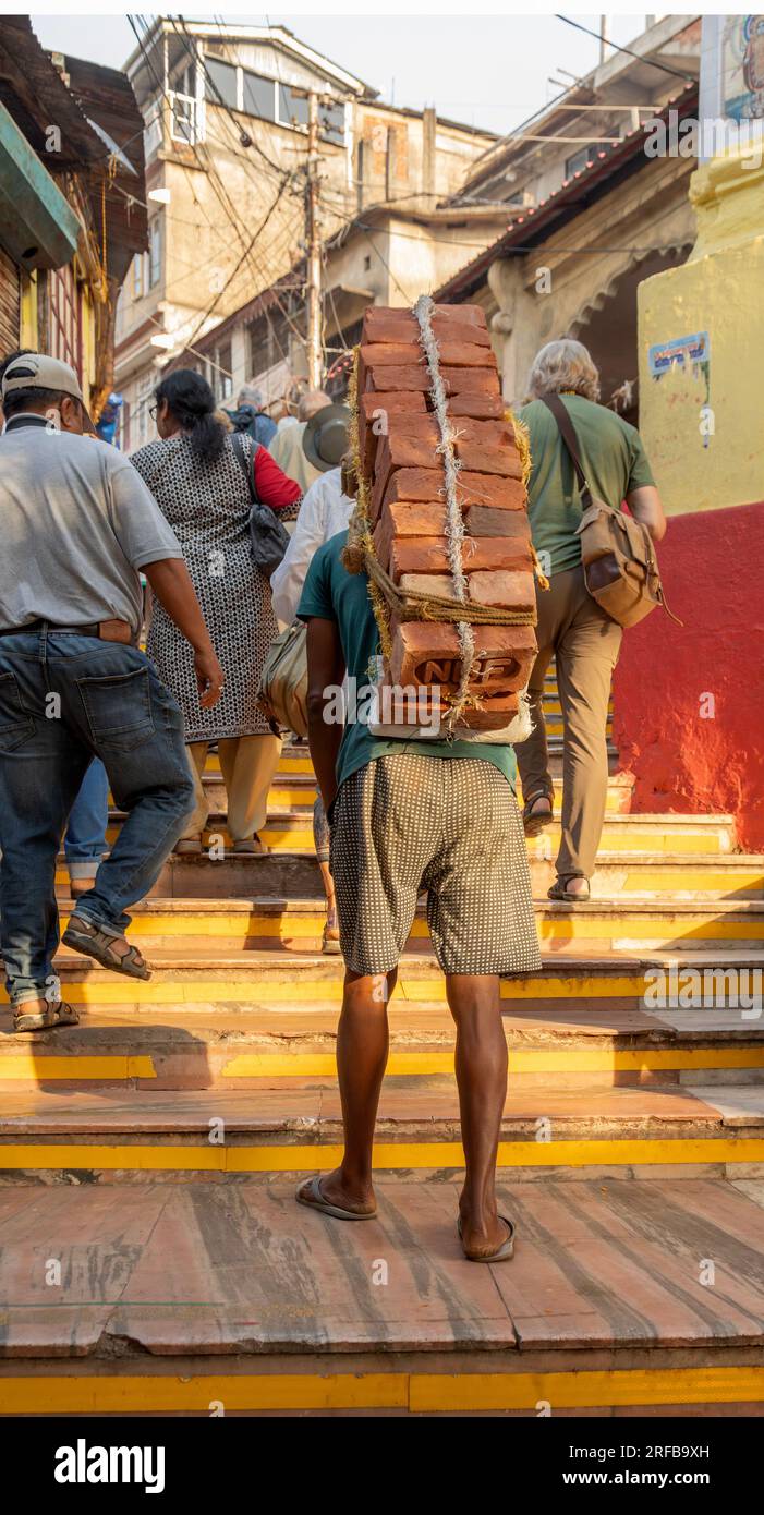 Un uomo che indossa infradito e porta un carico di mattoni sulla schiena, scalando i gradini verso il tempio di Kamakhya, Guwahati, dietro pellegrini e turisti. Foto Stock