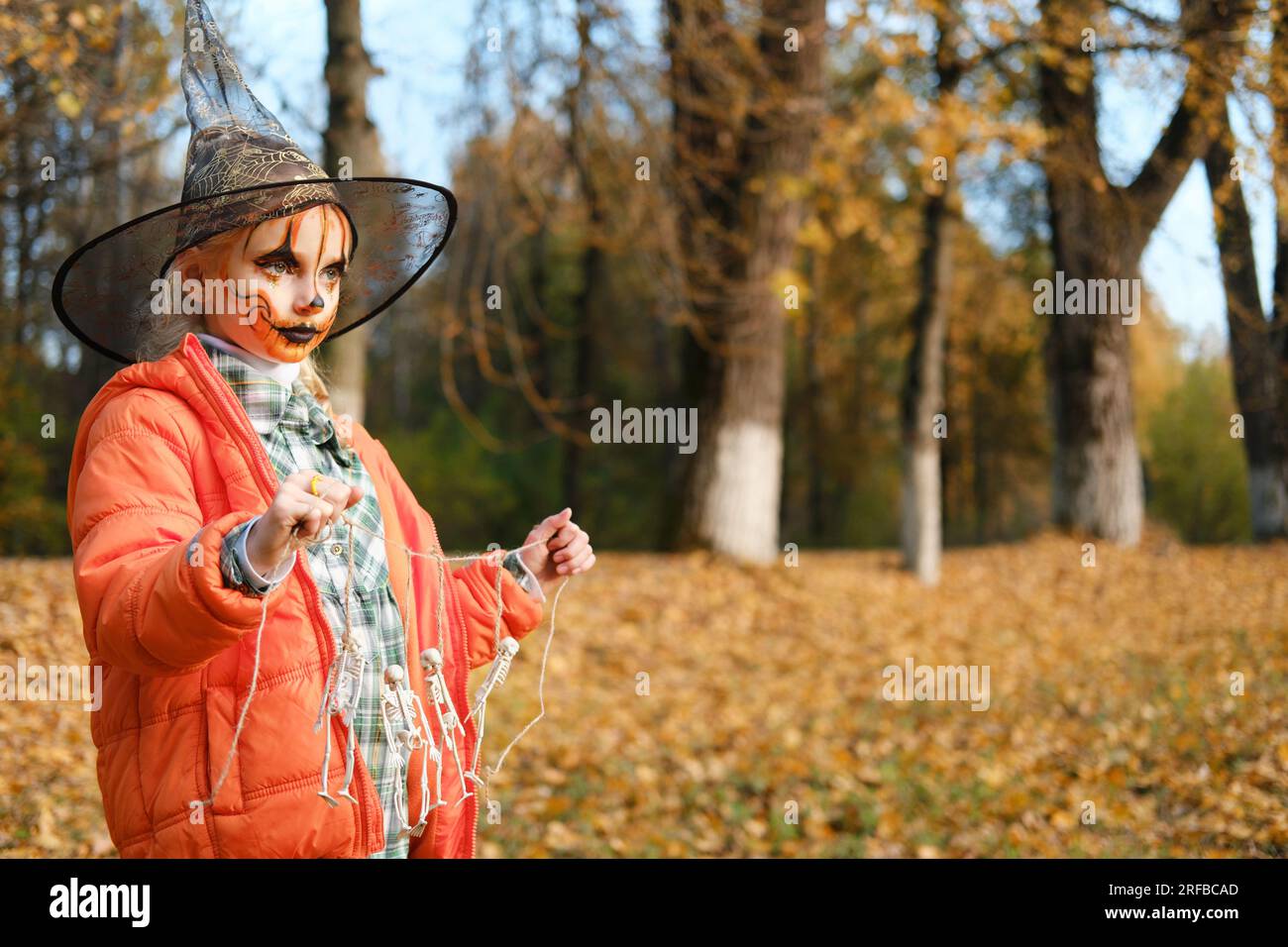 Una bambina con una giacca arancione, un cappello da strega e un trucco sul viso tiene in mano una decorazione di scheletri. Festa di Halloween. Horiz Foto Stock