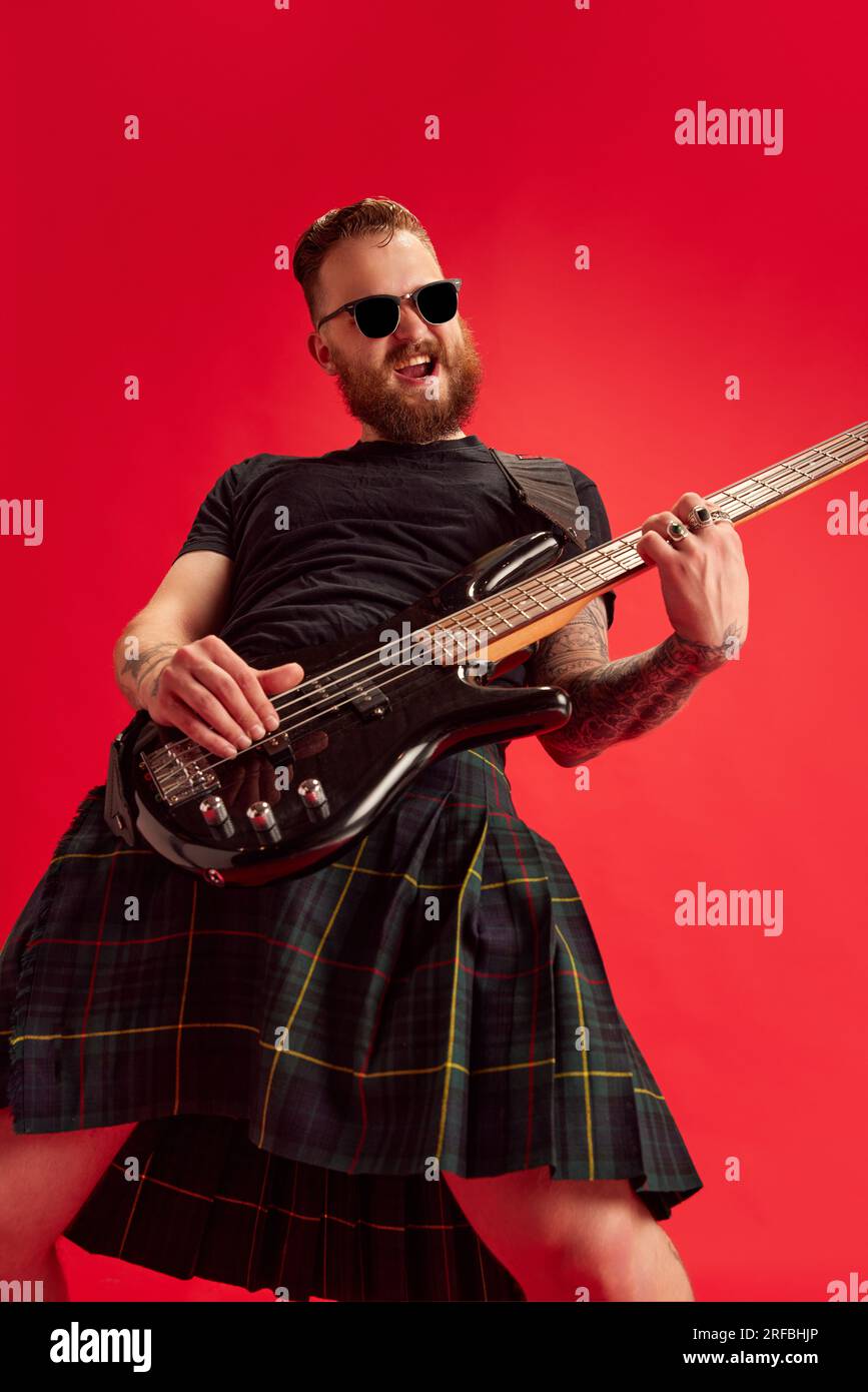 Concerto rock and roll. Uomo barbuto con occhiali da sole e gonna, kilt che suona la chitarra sullo sfondo rosso dello studio Foto Stock