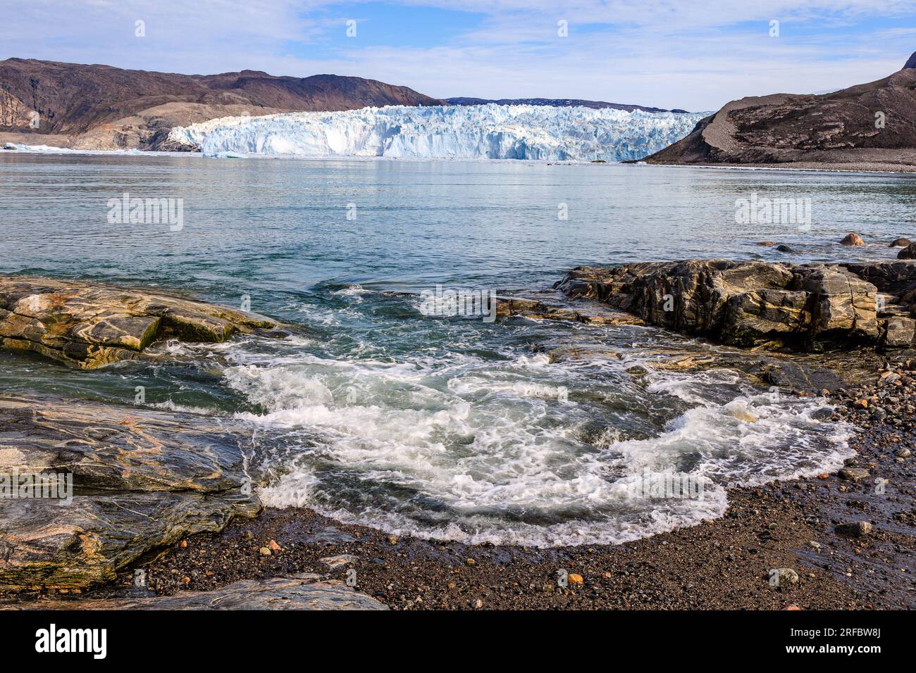paesaggio marino di un'onda che si rompe dolcemente su basse rocce con l'enorme ghiacciaio eqip sermia sullo sfondo Foto Stock