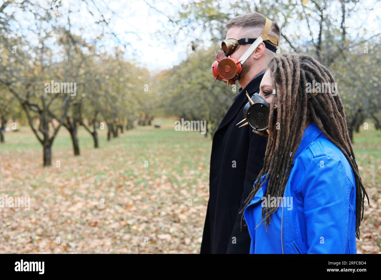 Una donna con dreadlock e una maschera a gas e un uomo con occhiali steampunk e una maschera a gas posa in un parco autunnale. Concetto di disastro ecologico. Orizzontale Foto Stock