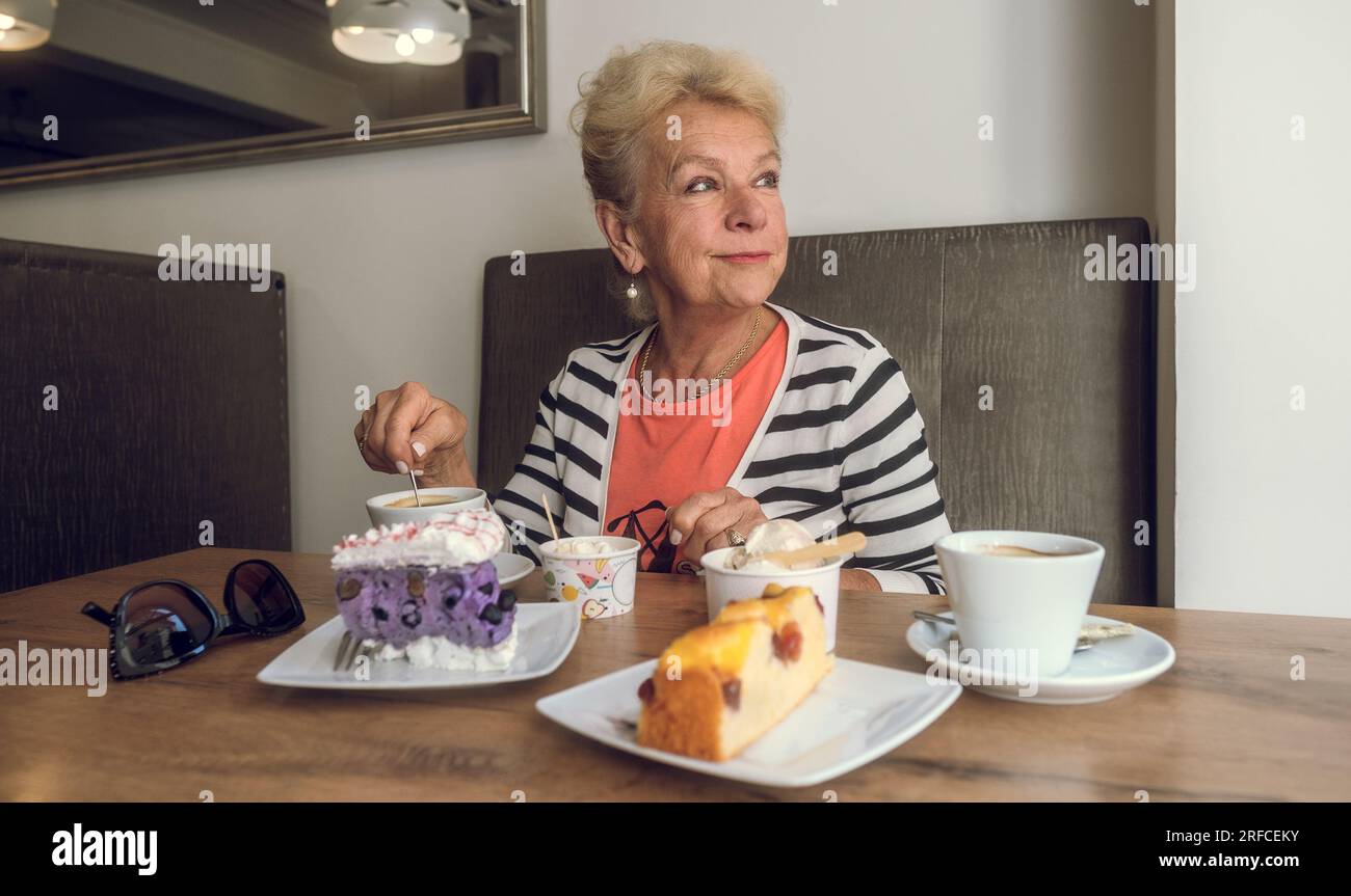 Donna anziana attraente e gioiosa che beve caffè mentre si siede in un bar. Ritratto di donna matura in una caffetteria. Torte colorate e caffè su un tavolo. Foto Stock