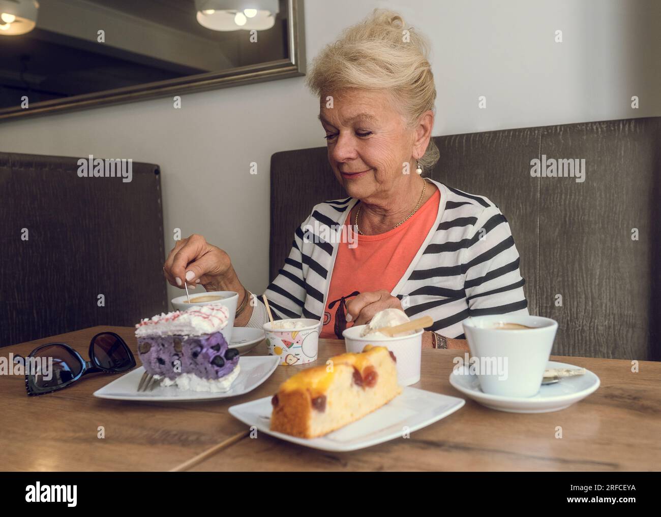 Donna anziana attraente e gioiosa che beve caffè mentre si siede in un bar. Ritratto di donna matura in una caffetteria. Torte colorate e caffè su un tavolo. Foto Stock