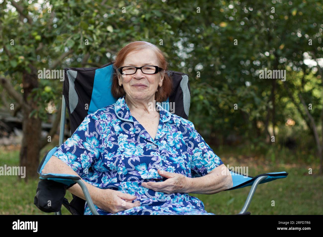 Ritratto della nonna anziana sorridente in giardino a casa. l'anziana si sta riposando seduto sulla sedia, guardando la macchina fotografica, il frutteto sullo sfondo. Somma Foto Stock