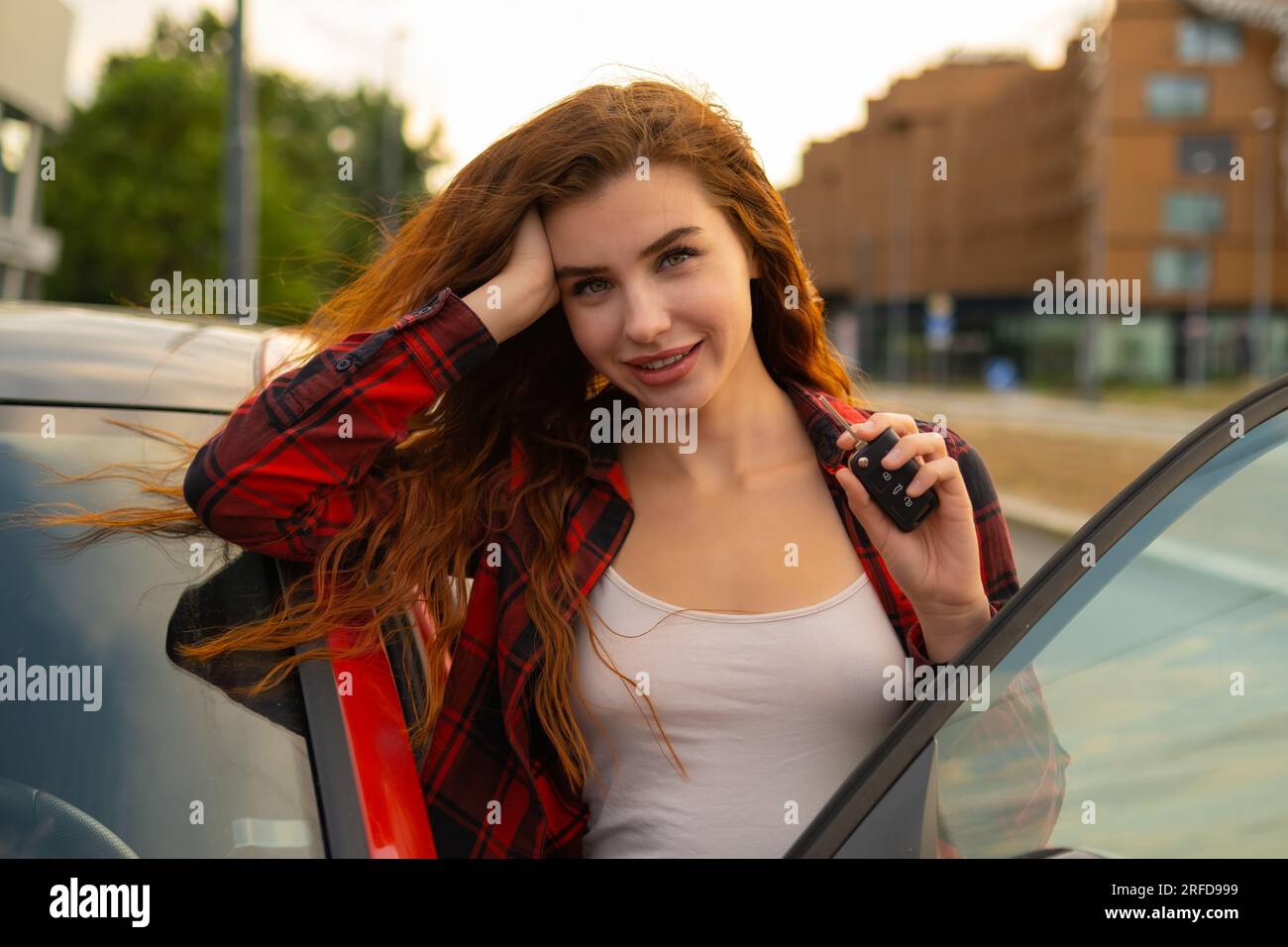 Con lucchetti rossi infuocati e un sorriso sorridente, la giovane donna che indossa una camicia a quadri casual posa accanto all'auto rossa e tiene le chiavi dell'auto in mano. Foto Stock