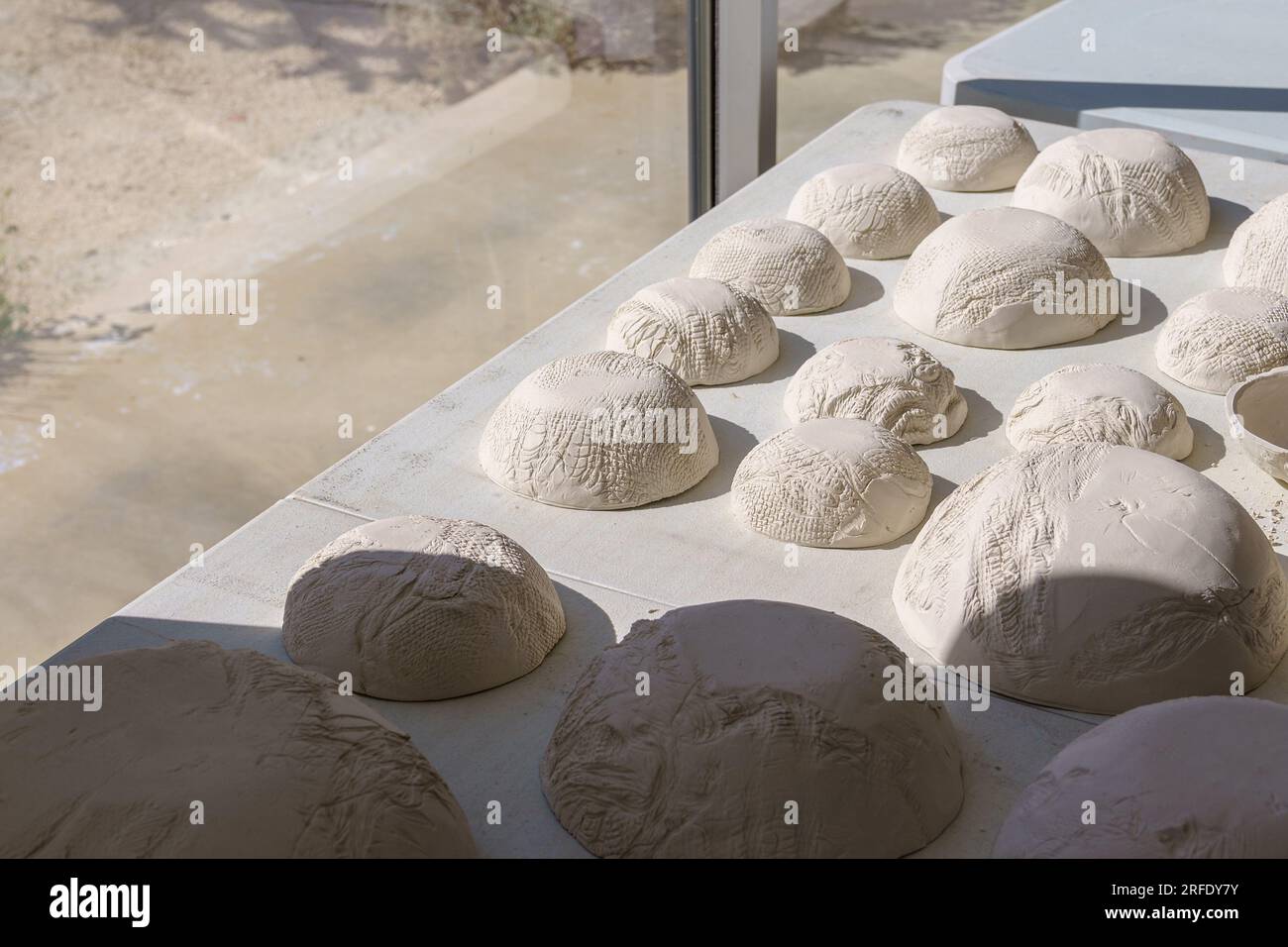Una collezione e una varietà di ciotole in argilla bianca cruda fatte a mano sedute al contrario sotto sotto la luce del sole accanto a una grande finestra Foto Stock