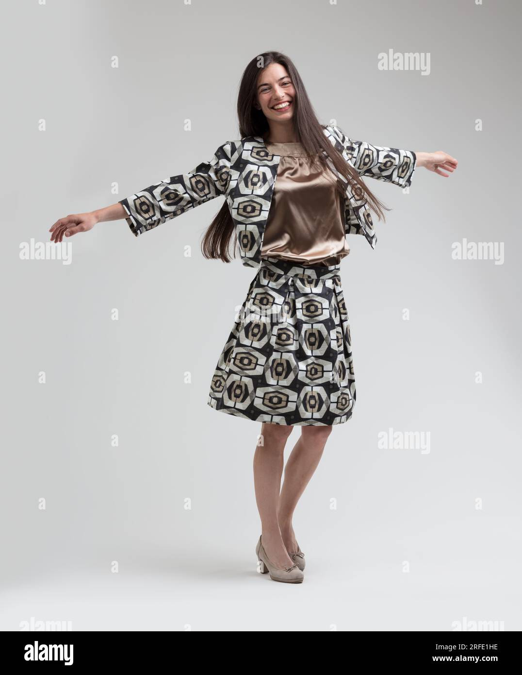 Modello gioioso con lunghi capelli giradischi, a braccio aperto, danzante in un abito sartoriale lungo il ginocchio, blusa color beige argento. Gira felicemente, ama la moda, wea Foto Stock