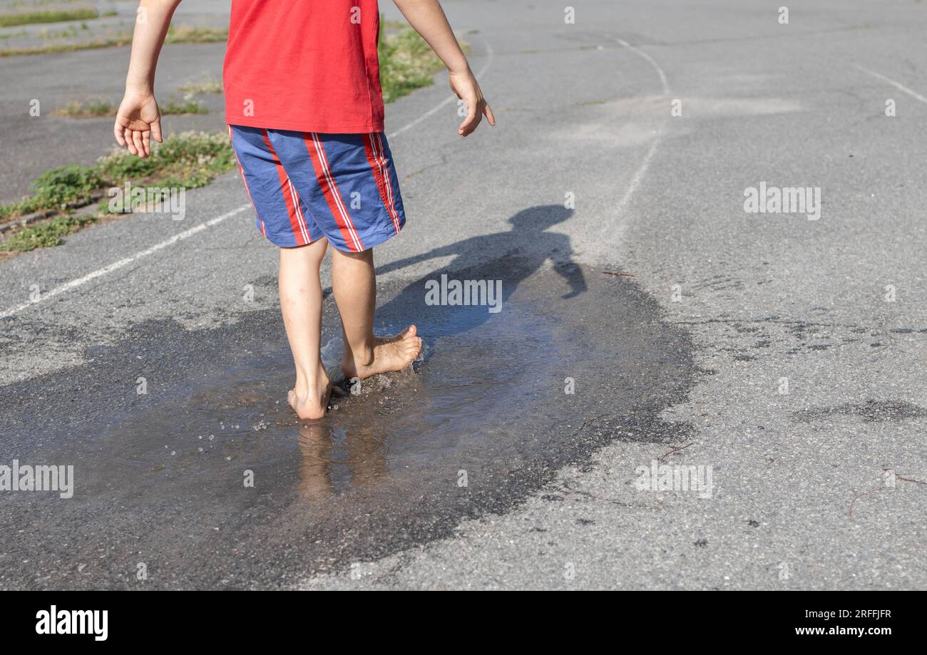 un bambino scalzo irriconoscibile cammina con gioia in una pozzanghera trasparente sul marciapiede dopo la pioggia. Cambiamenti del tempo in estate. Interessante divertente chi Foto Stock