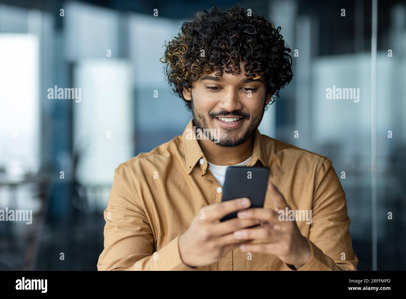 Primo piano di un giovane uomo d'affari che utilizza un'applicazione online al telefono, uomo in ufficio che legge buone notizie dallo smartphone, uomo latino-americano sorridente. Foto Stock
