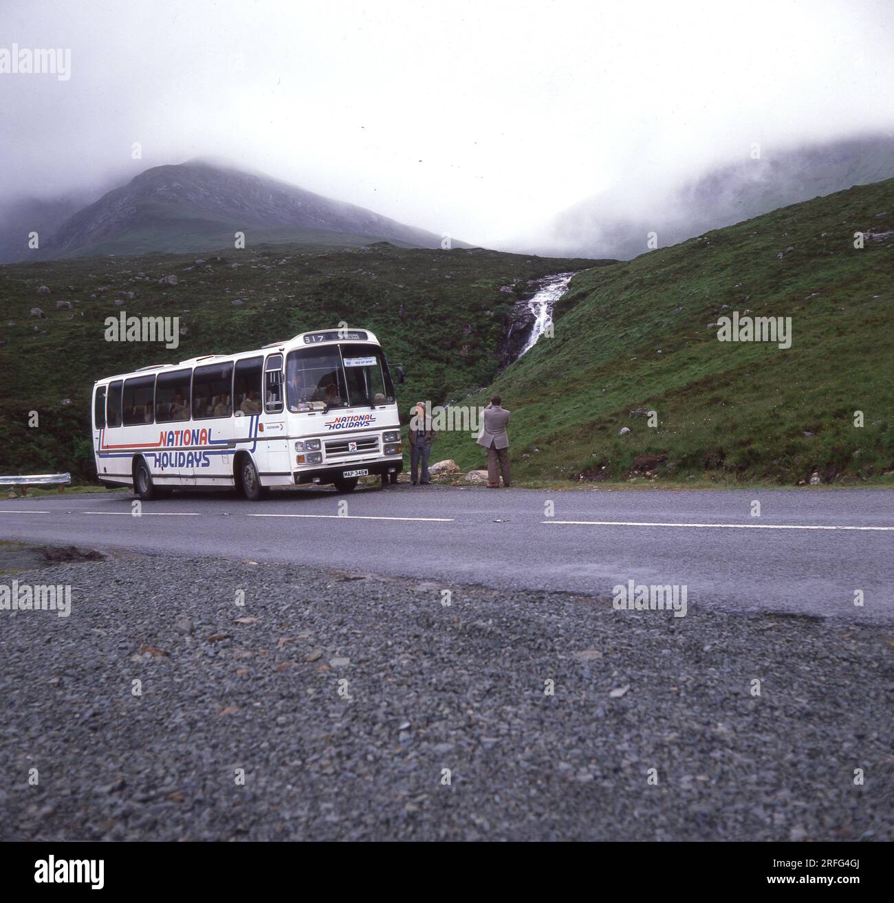 Fine degli anni '1970, storico, un pullman delle feste nazionali parcheggiato su una strada di montagna sull'Isola di Skye, Scottish Highlands, Scozia, Regno Unito. Un viaggiatore di sesso maschile fuori dal pullman che scatta una foto della valle nebbiosa. National Holidays era il ramo turistico della National Bus Company, una compagnia di autobus nazionalizzata, che era stata costituita nel 1969. La NBC è stata rinominata National Express nel 1974, con il nuovo marchio di strisce blu e bianche introdotto nel 1978. A metà degli anni '80, la National Bus Company fu smantellata, con National Holidays venduta a Pleasurama nel 1986. Foto Stock