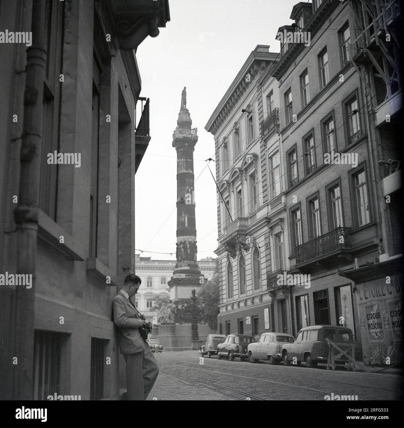 Anni '1950, storico, un uomo con una cinepresa in piedi in una strada lastricata con binari del tram a Parigi, in Francia, con il monumento alla guerra, le colonne Vendome viste sullo sfondo. Un poster sbiadito per le Cirque de Moscou sul muro di fronte a un edificio. Foto Stock