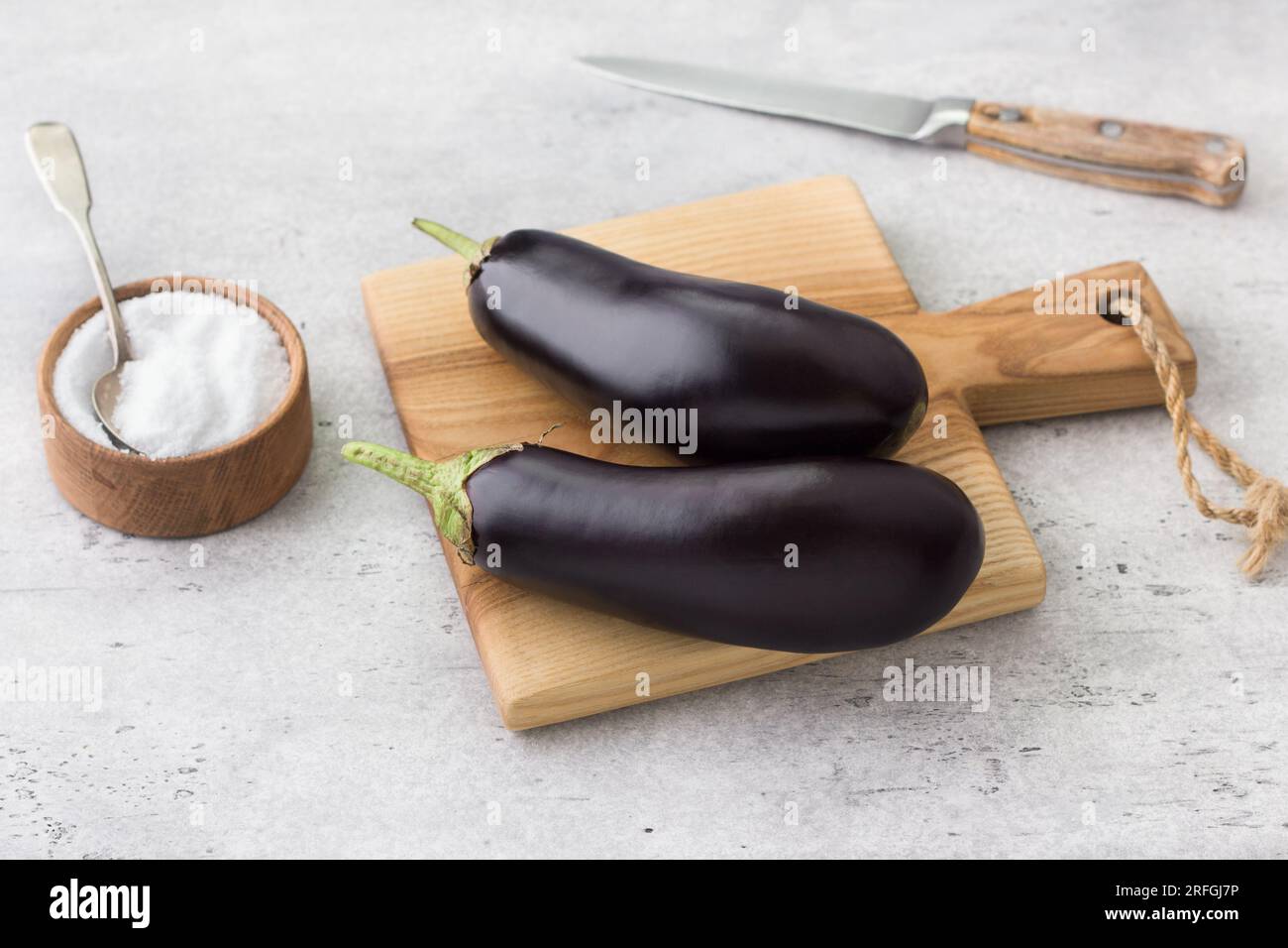 Melanzana fresca su un asse di legno, una ciotola di sale e un coltello su uno sfondo grigio ruvido. Cucinare deliziosi piatti vegani. Foto Stock