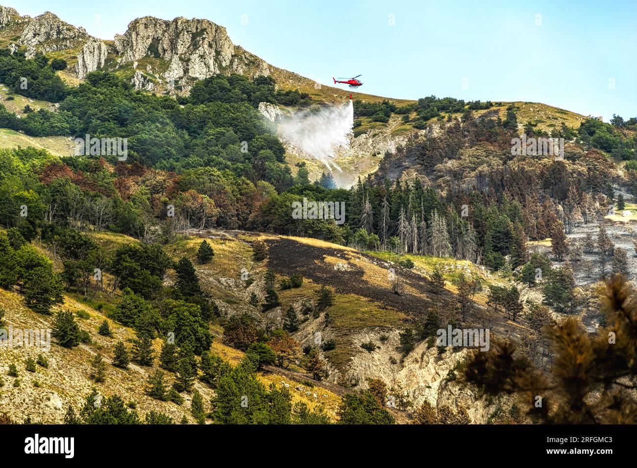 Scena drammatica con fiamme e fumo provenienti da un incendio boschivo. L'elicottero della protezione civile combatte le fiamme con l'acqua Monte Morrone, Parco Nazionale della Maiella Foto Stock