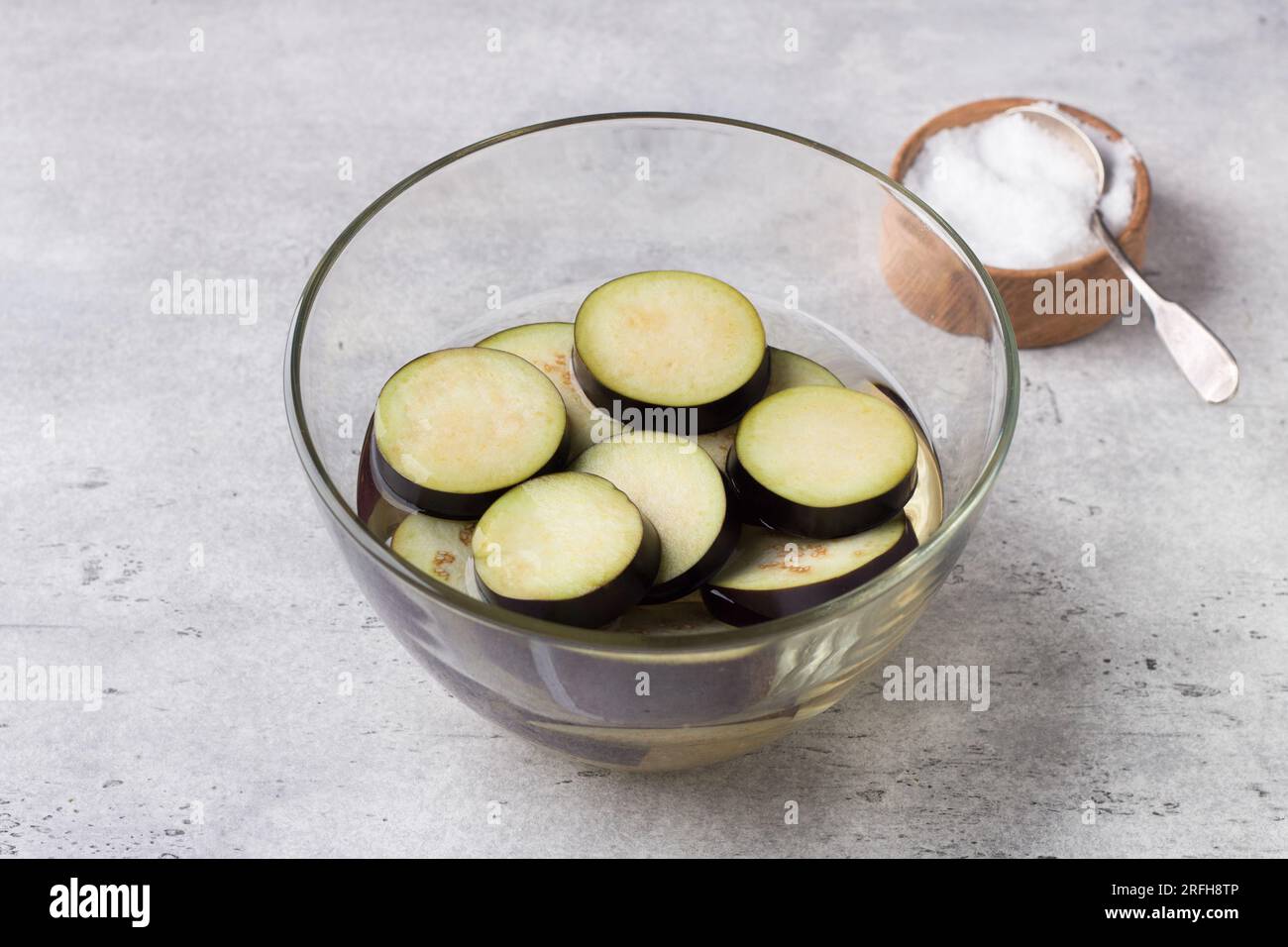 Immergere le melanzane in acqua salata in una ciotola di vetro su uno sfondo grigio ruvido. Lavorazione delle melanzane prima della cottura, eliminando l'amaro. Foto Stock