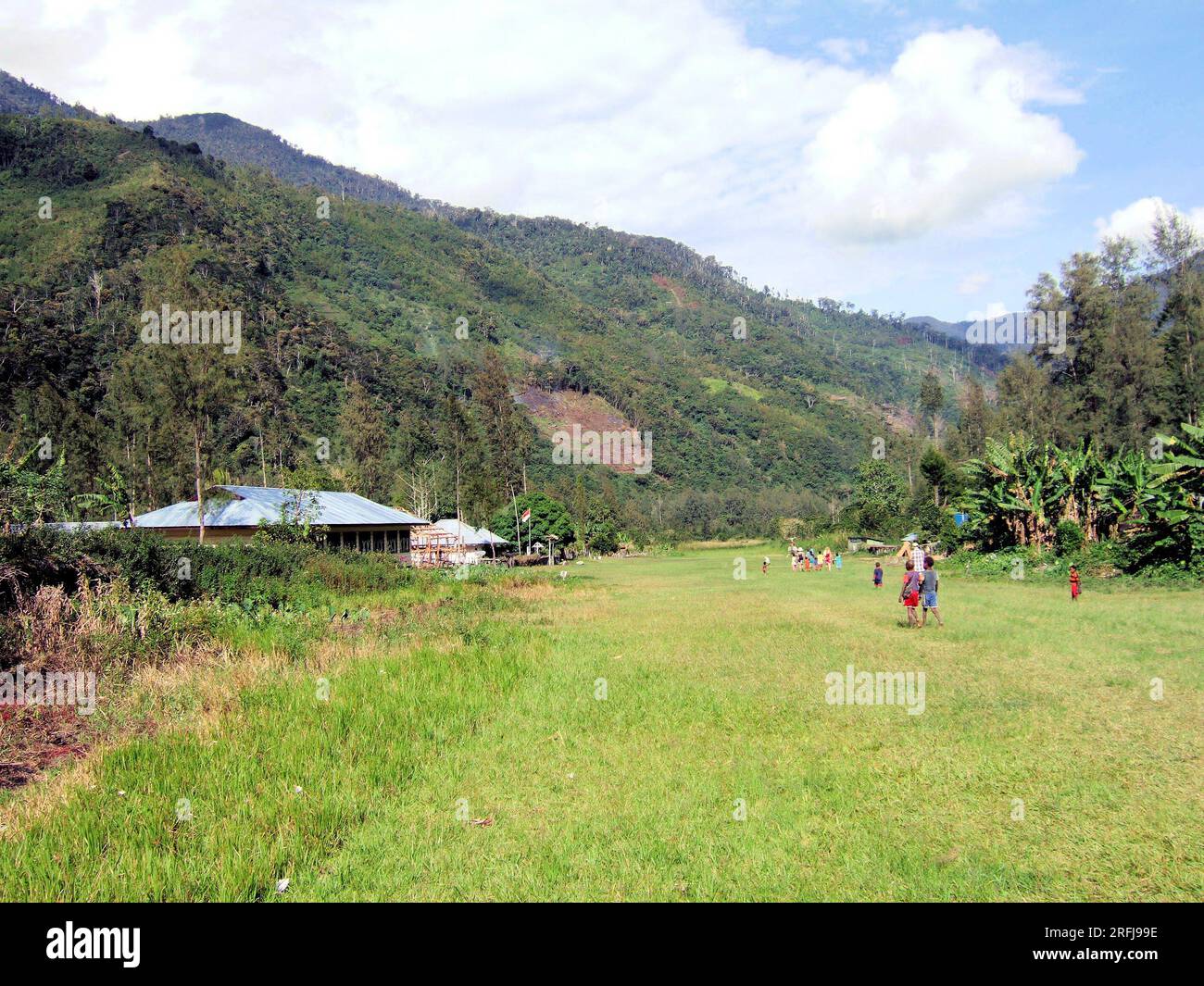 Papuani che camminano in lontananza su una piccola pista di atterraggio, con edifici e montagne sullo sfondo. Foto Stock