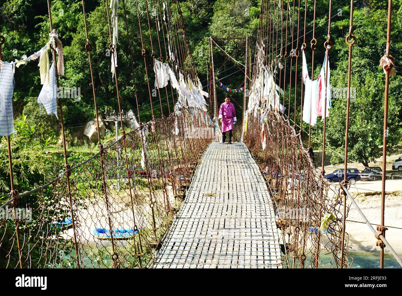 Un uomo bhutanese che indossa il tradizionale Gho cammina attraverso un ponte sospeso nel Bhutan rurale. Le bandiere di preghiera sbiadite si appendono ai cavi di supporto in metallo. Foto Stock