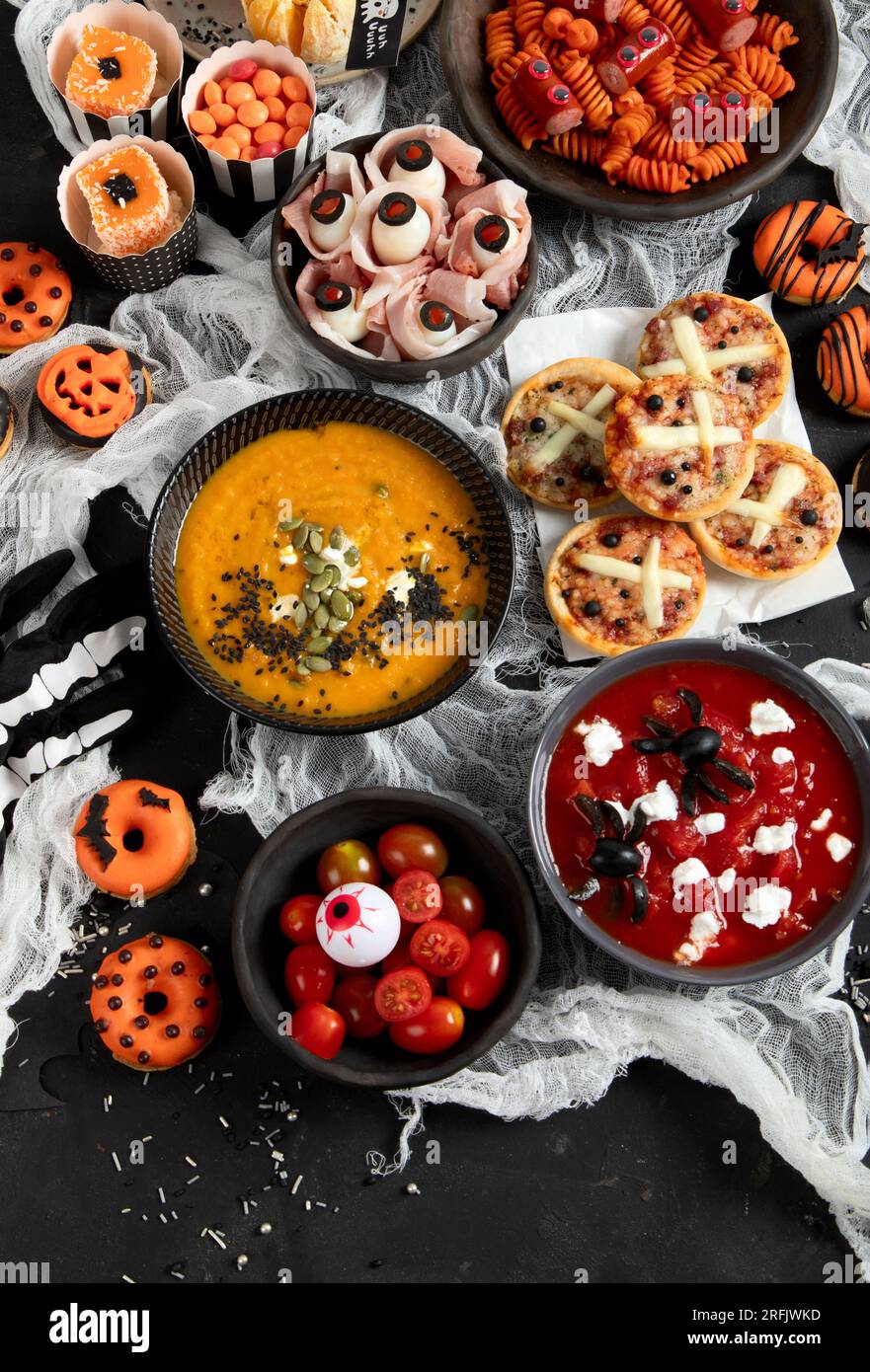 Divertente tavolo da festa di Halloween. Pizza, zuppa di zucca, caramelle, spaghetti oculari, spuntini e ciambelle su sfondo nero. Vista dall'alto. Foto Stock