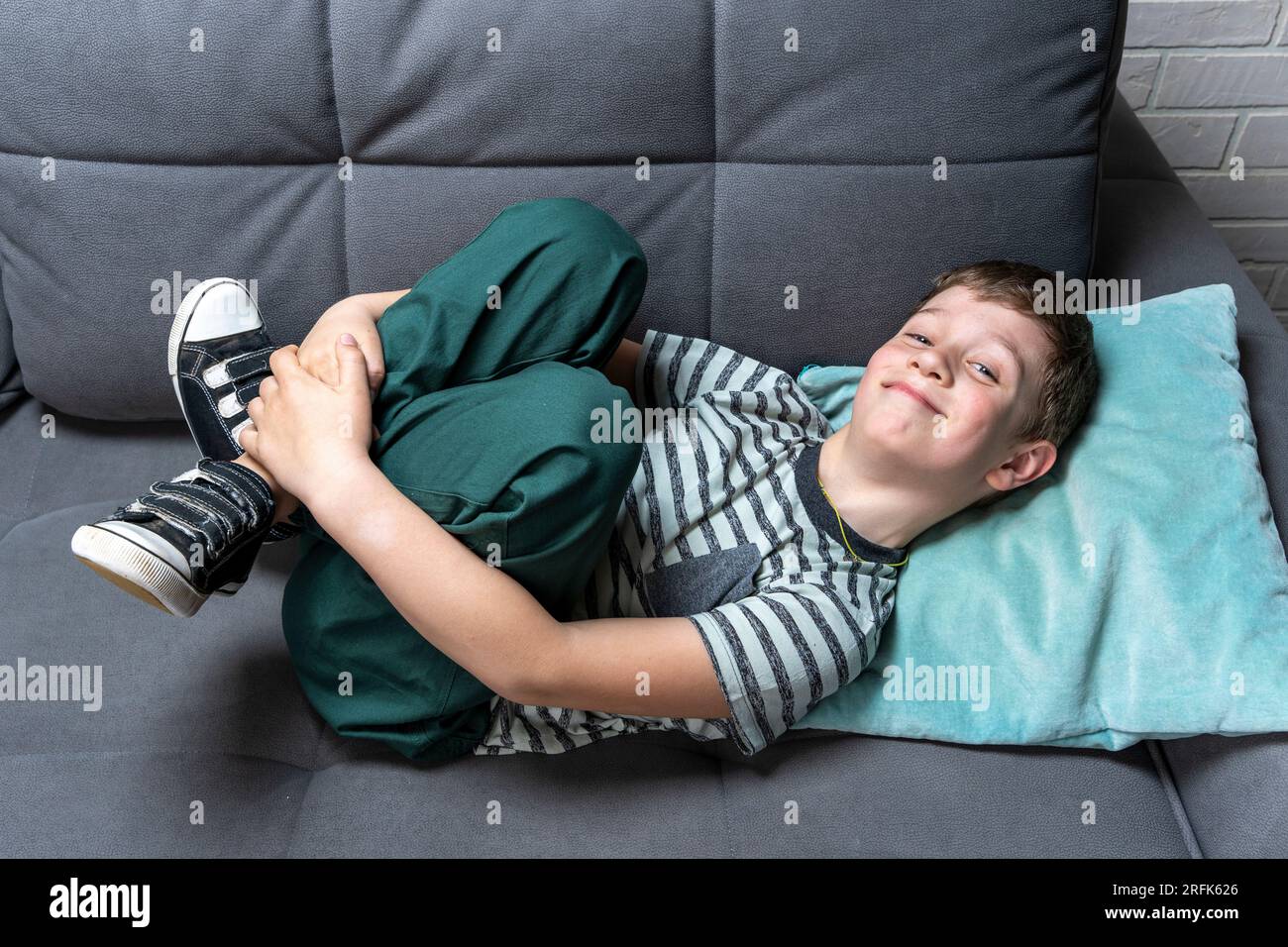 Un allegro ragazzo caucasico in età scolare è sdraiato sul divano e ingannato sorridendo. Attività ricreative e disattenzione dei bambini Foto Stock
