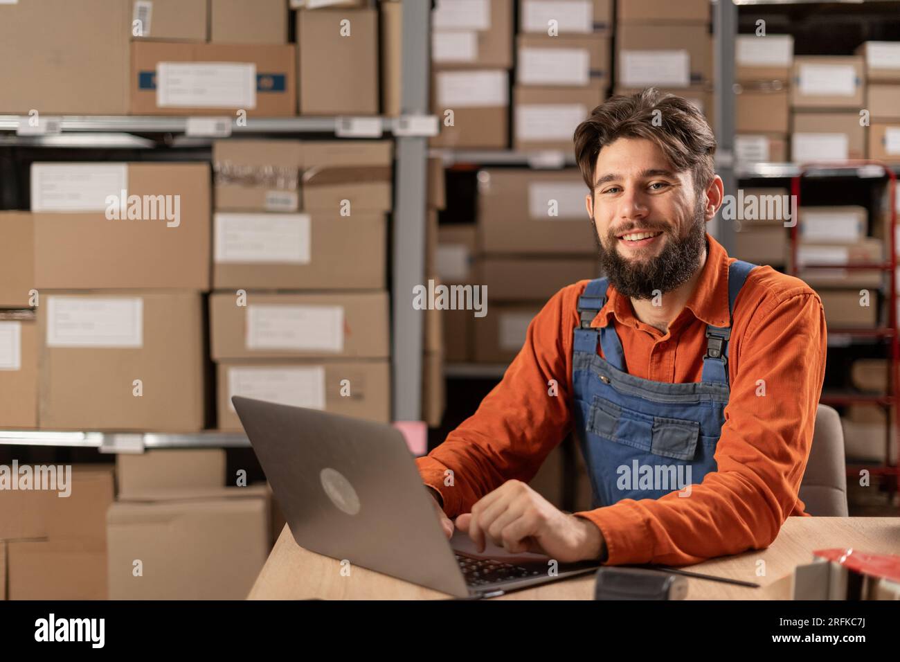 Addetto al magazzino con notebook che immette i dati di inventario nel database, sorride e guarda la fotocamera Foto Stock