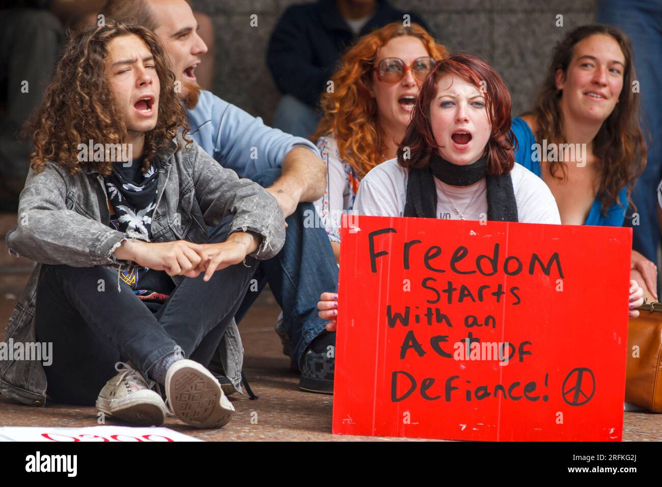 Un gruppo di manifestanti seduti sul marciapiede cantano e tengono uno striscione che afferma che la libertà inizia con un atto di sfida Foto Stock