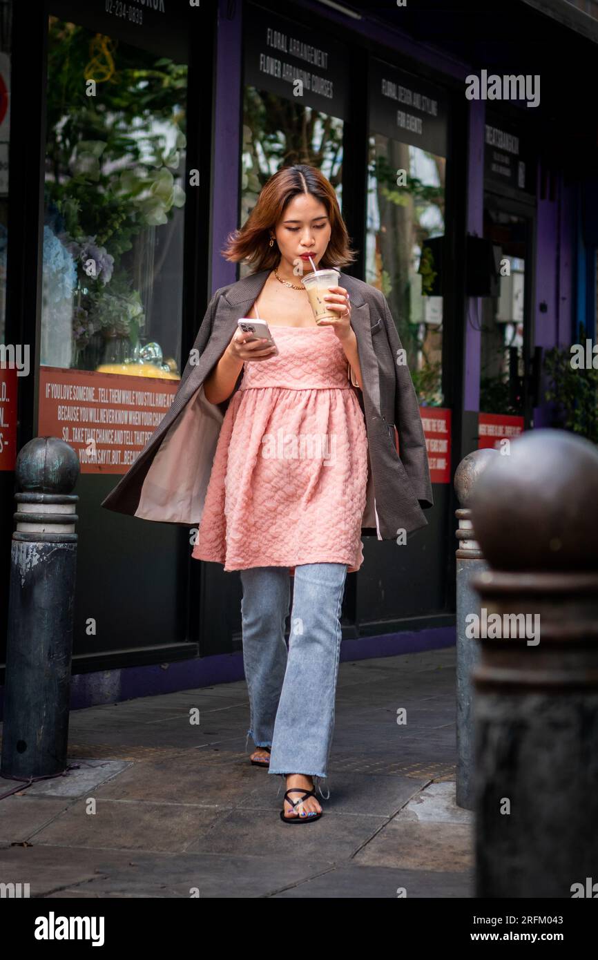 Una giovane ragazza thailandese sorseggia il caffè e controlla il suo telefono mentre scende nella sala Daeng Rd. Bangkok Thailandia. Foto Stock