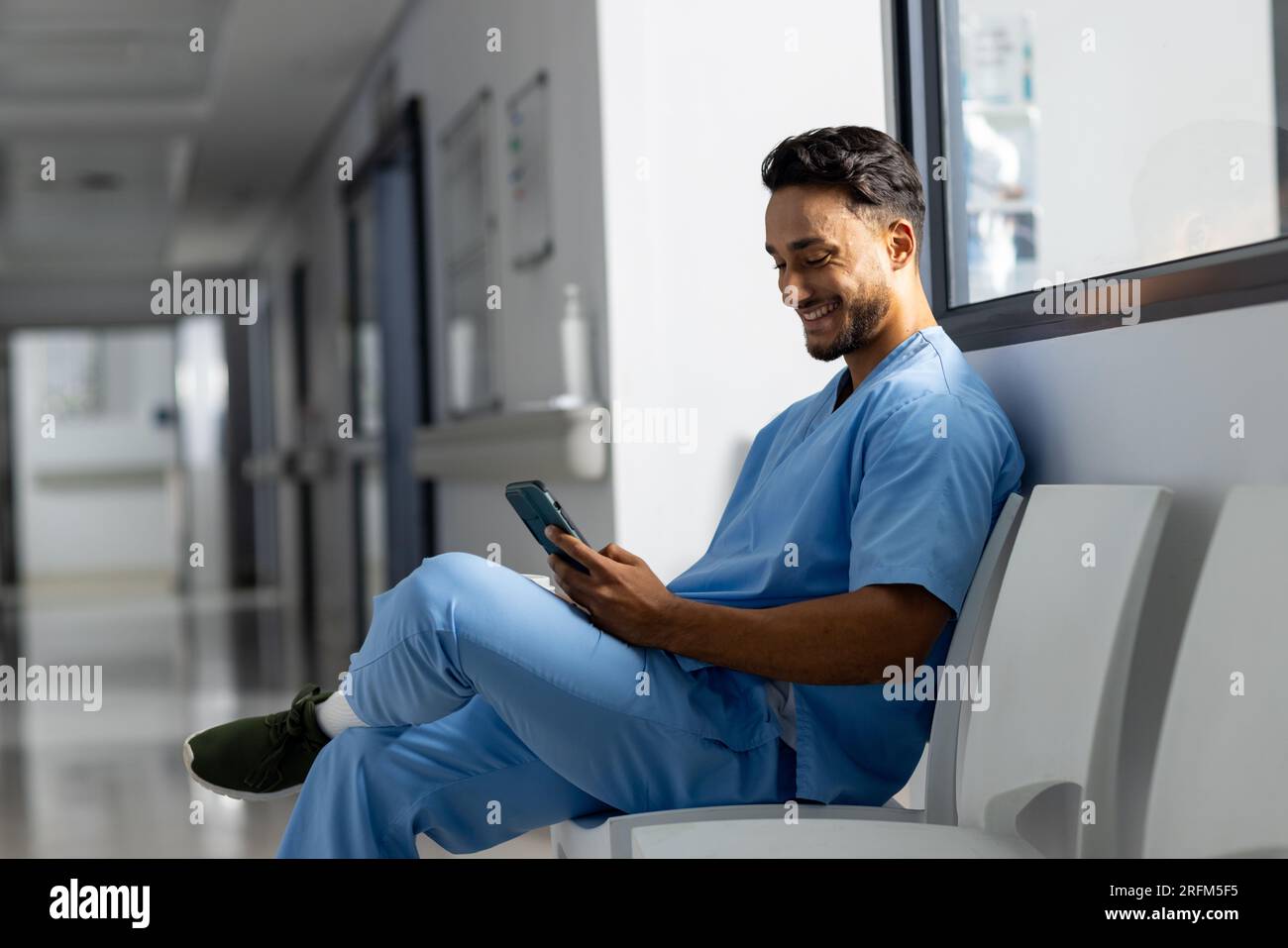 Medico maschio birazziale che indossa scrub utilizzando smartphone in corridoio all'ospedale Foto Stock