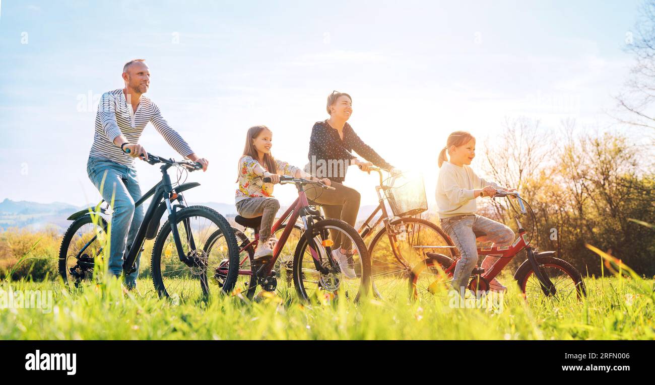 Sorridente padre e madre con due figlie durante l'estate in bicicletta all'aperto. Si godono insieme sul prato verde alto prato. Felice parenthoo Foto Stock