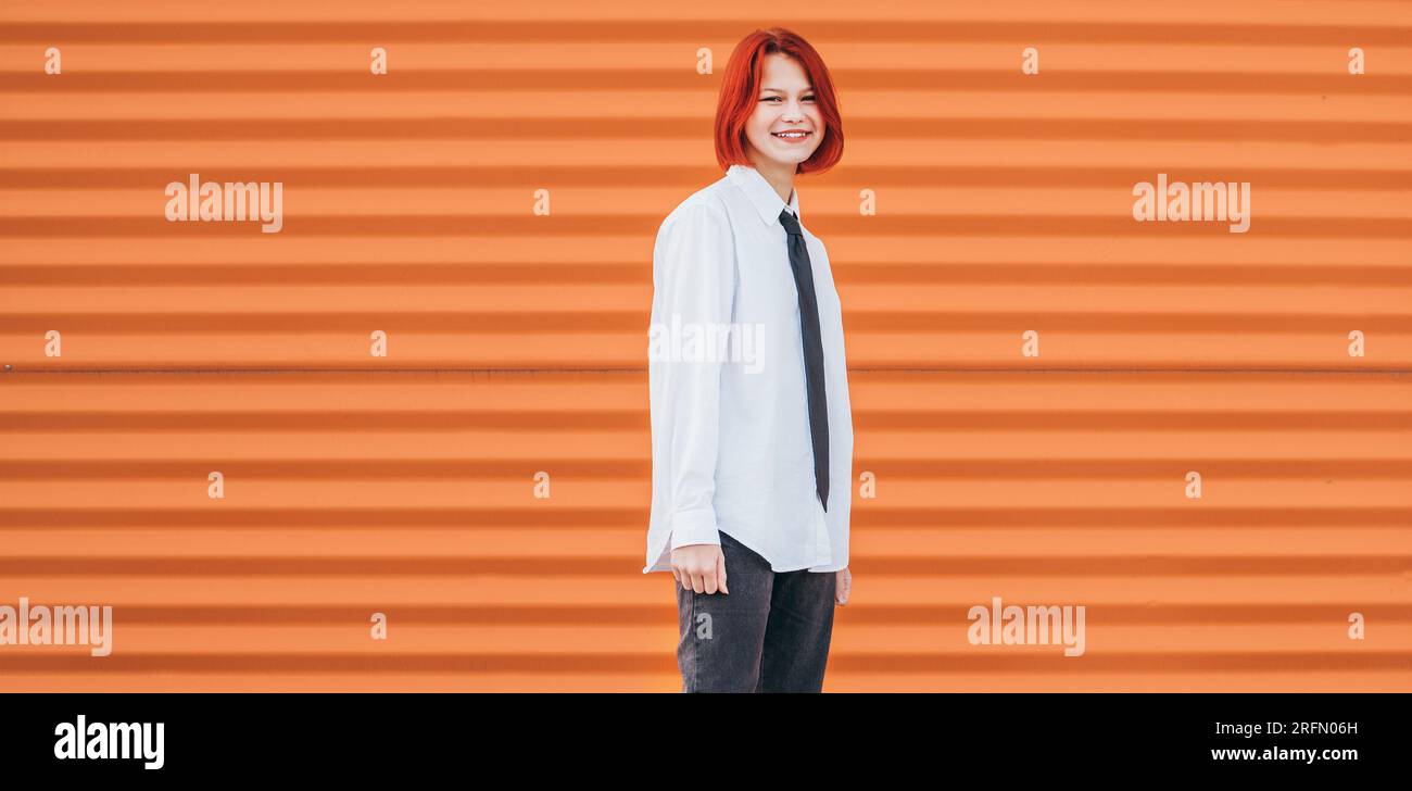 Ritratto di un'adolescente sorridente con capelli tinti di rosso con camicia bianca e cravatta in piedi vicino a sfondo arancione sulla zampa pedonale Foto Stock