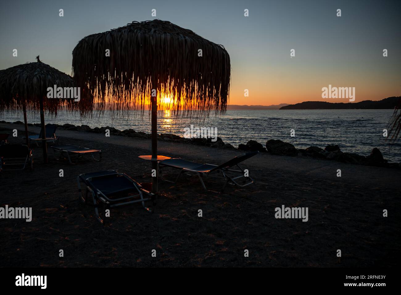 All'alba del resort, i raggi del sole sgorgano attraverso l'ombrellone dalle foglie di palma. Sagome di sdraio, ombrelloni e molo. Foto Stock