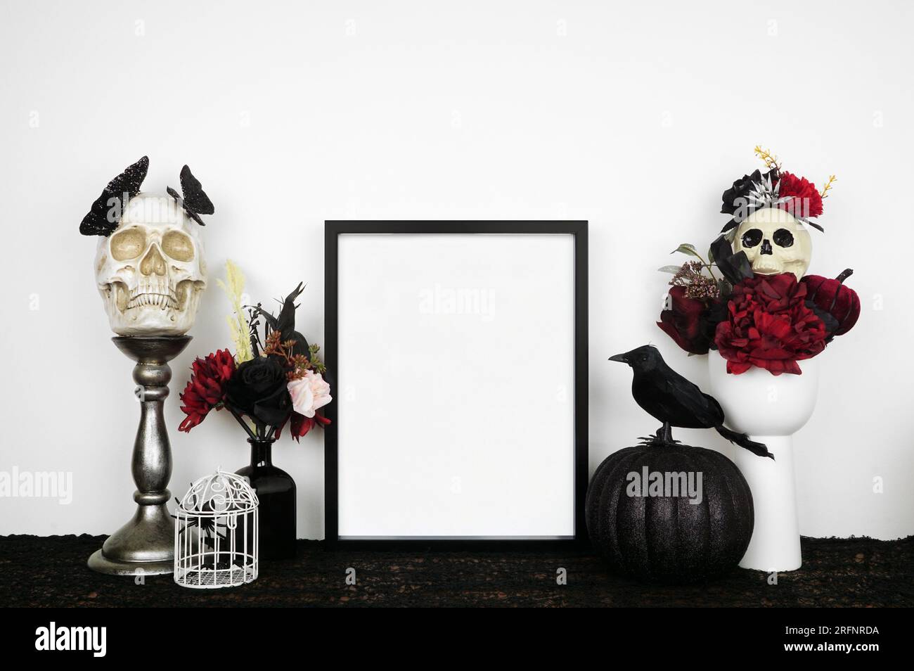 Halloween gothic Romance Mock-up. Cornice nera su uno scaffale nero con fiori rossi e neri, teschi e zucca. Cornice verticale contro una parete bianca. Co Foto Stock