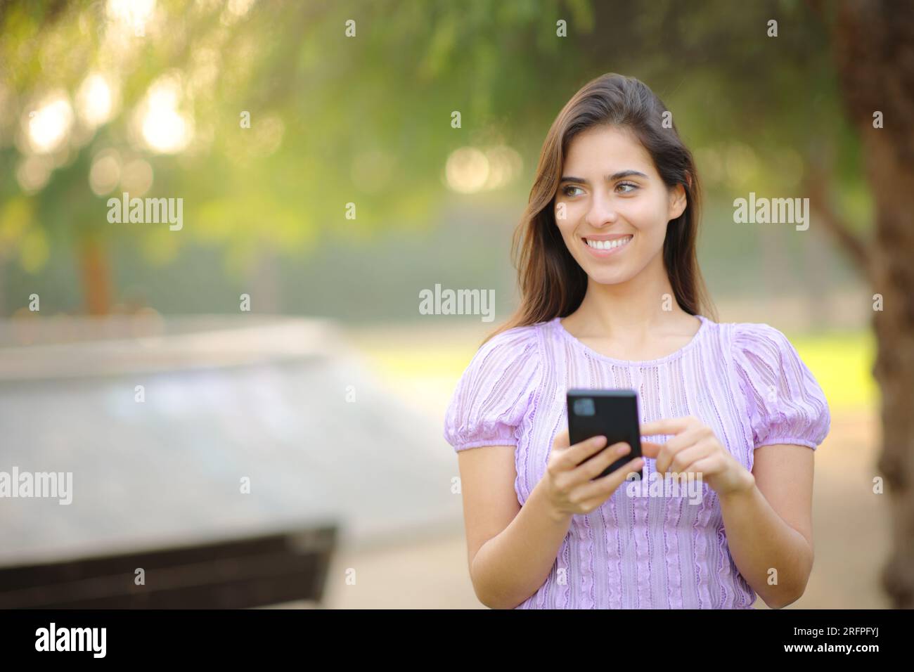 Ritratto frontale di una donna felice che usa il telefono guardando una passeggiata laterale in un parco Foto Stock