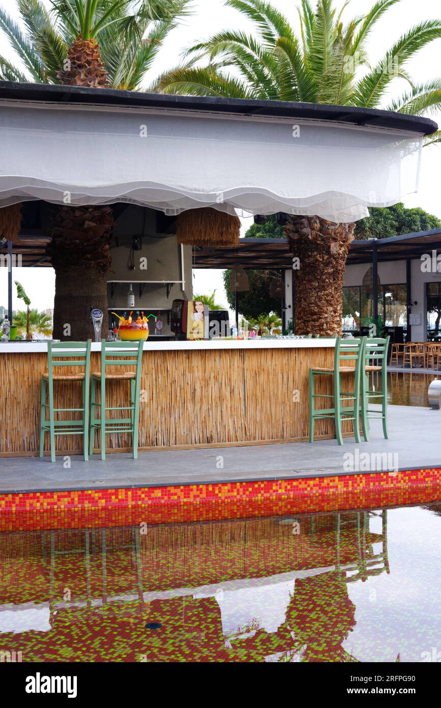 Tropic bar vicino alla piscina in un giorno d'estate con palme sullo sfondo. Riflesso sull'acqua e sulle sedie. Foto Stock