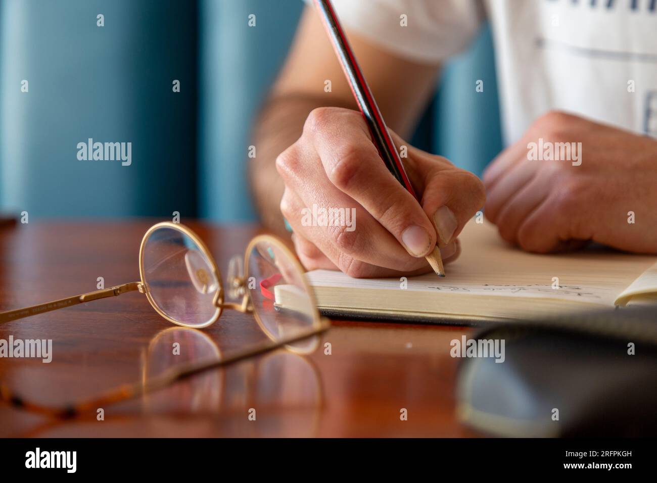 Un dettaglio di uno scrittore con gli occhiali e la matita che scrive su un giornale. Prendere appunti con la matita su un notebook con gli occhiali appoggiati. Foto Stock