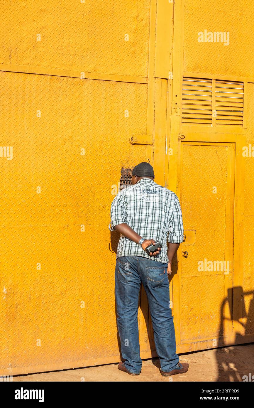 Uomo che discute attraverso un wicket perforato in una porta di metallo giallo intenso. Jinja, Uganda. Foto Stock