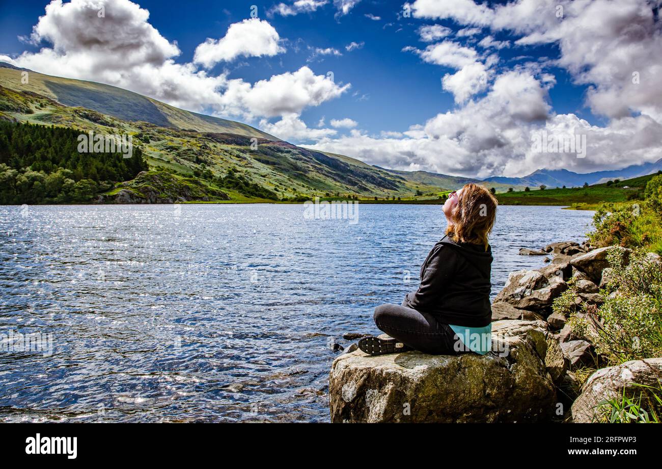 Donna adulta/anziana sedeva su una roccia accanto a un lago in posizione di meditazione yoga a gambe incrociate, guardando verso il sole circondato da montagne Foto Stock